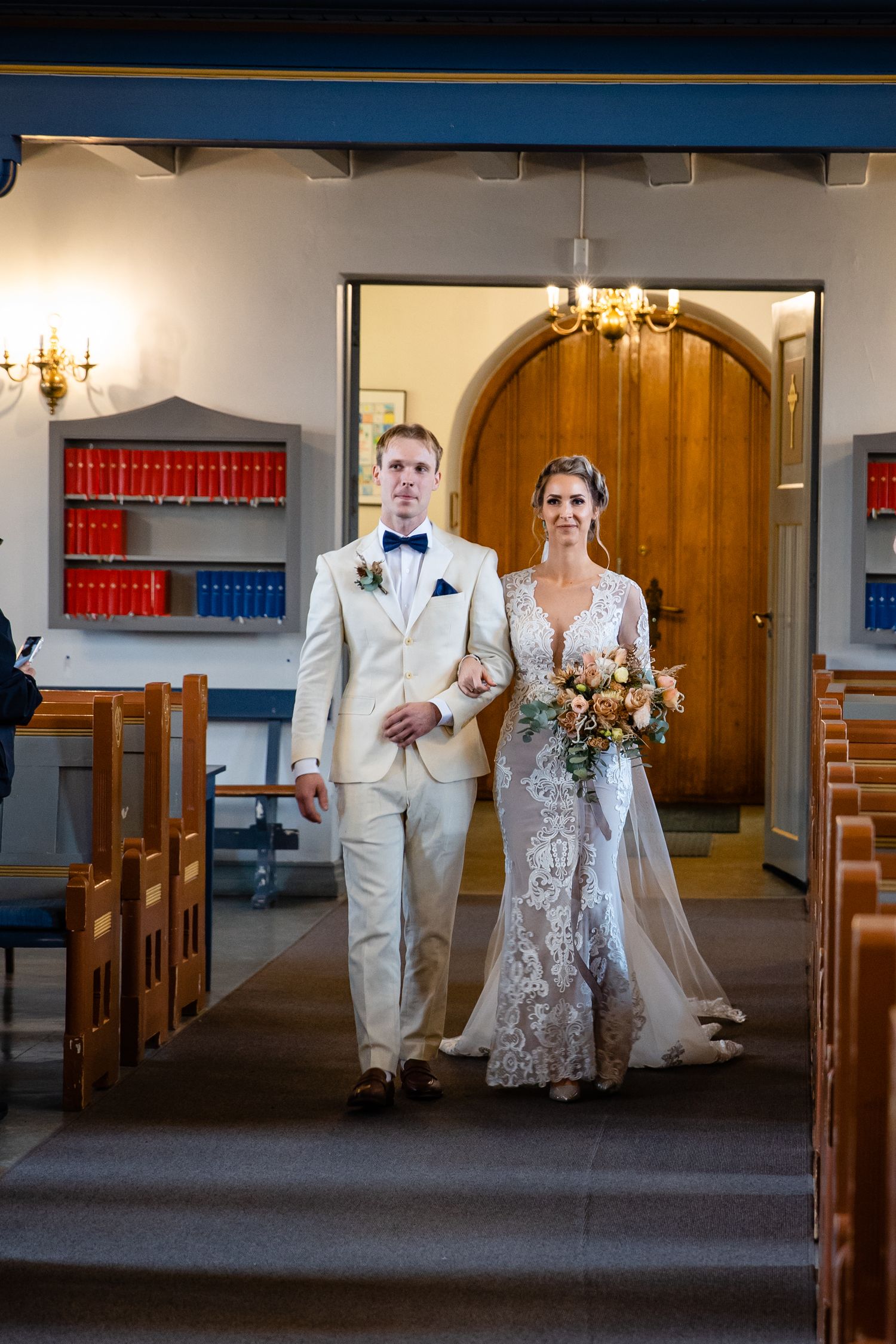 A couple poses in formal wedding attire inside a church with wooden pews and blue walls.