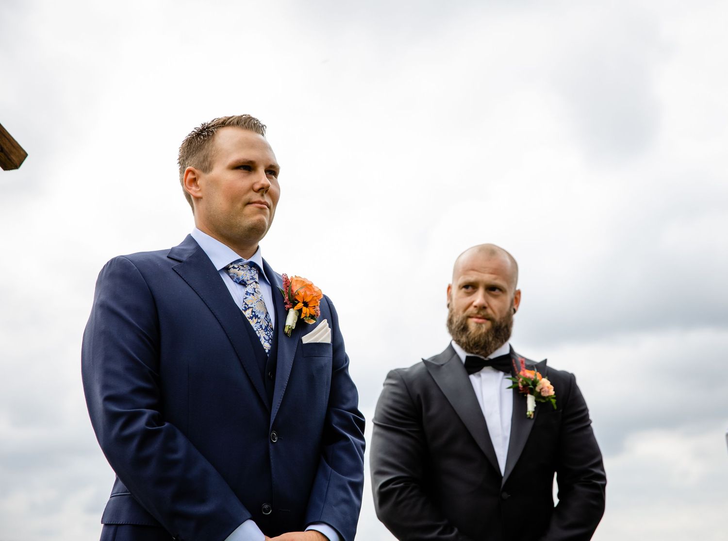 Groomsmen in navy suits stand together against cloudy sky during outdoor wedding ceremony.