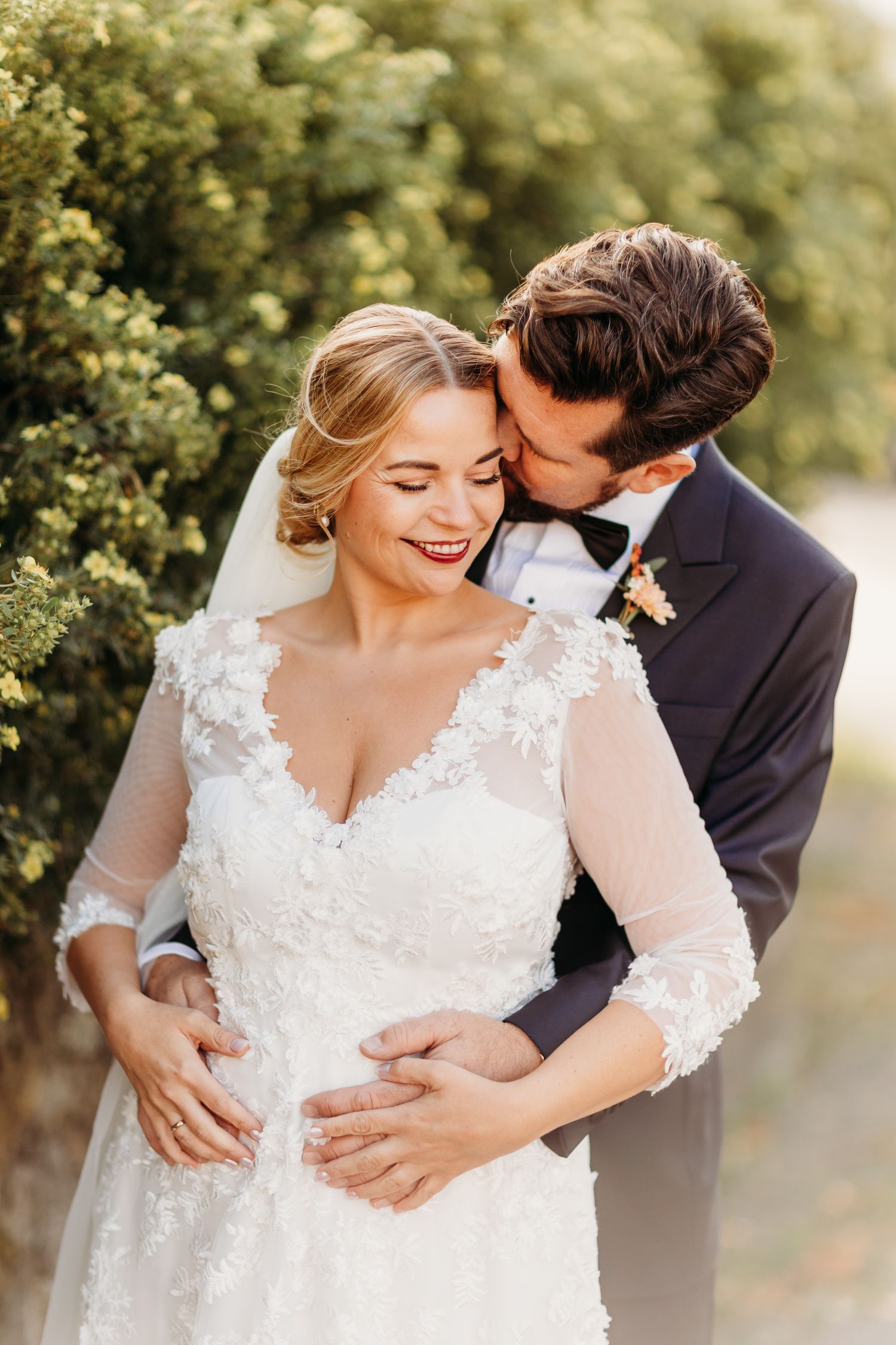 Romantic wedding portrait of couple embracing outdoors with bride in lace dress and groom in tuxedo.