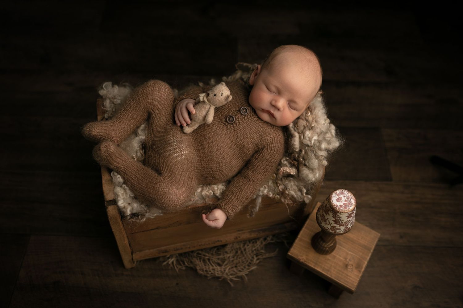 Nyfødtfotografering Moss: En sovende nyfødt i strikket drakt ligger trygt i en rustikk seng med en liten bamse, fotografert i studio i Våler