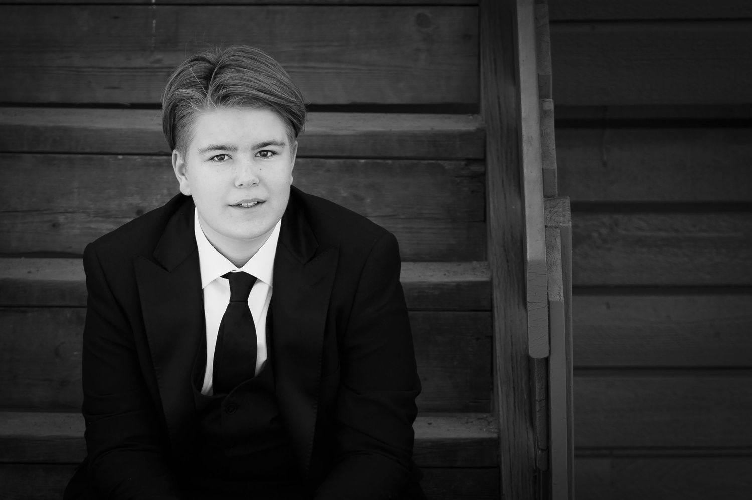 Monochrome portrait of person in black suit and tie against wooden wall background.
