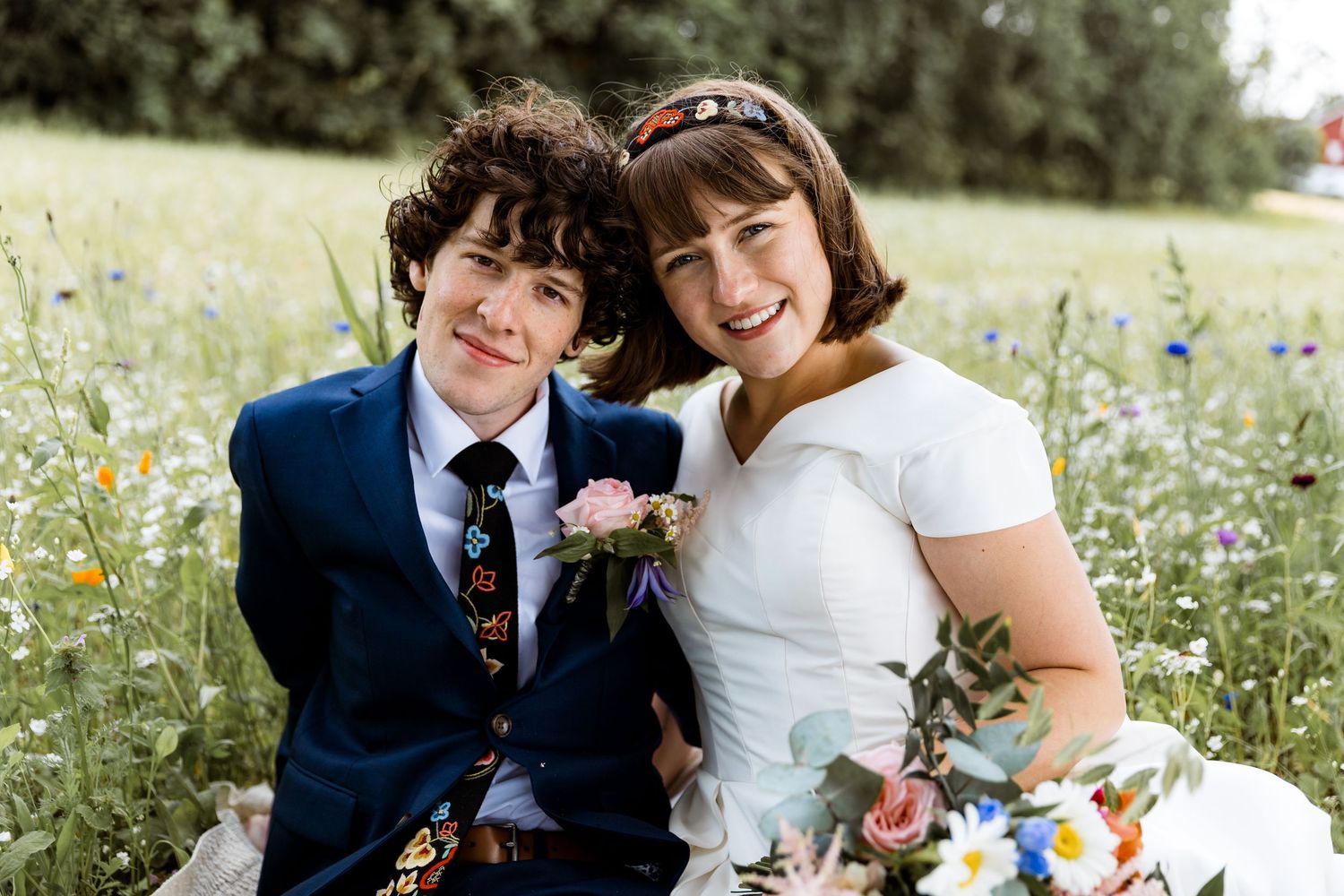 A joyful wedding couple sitting together in a wildflower meadow wearing blue and white attire.