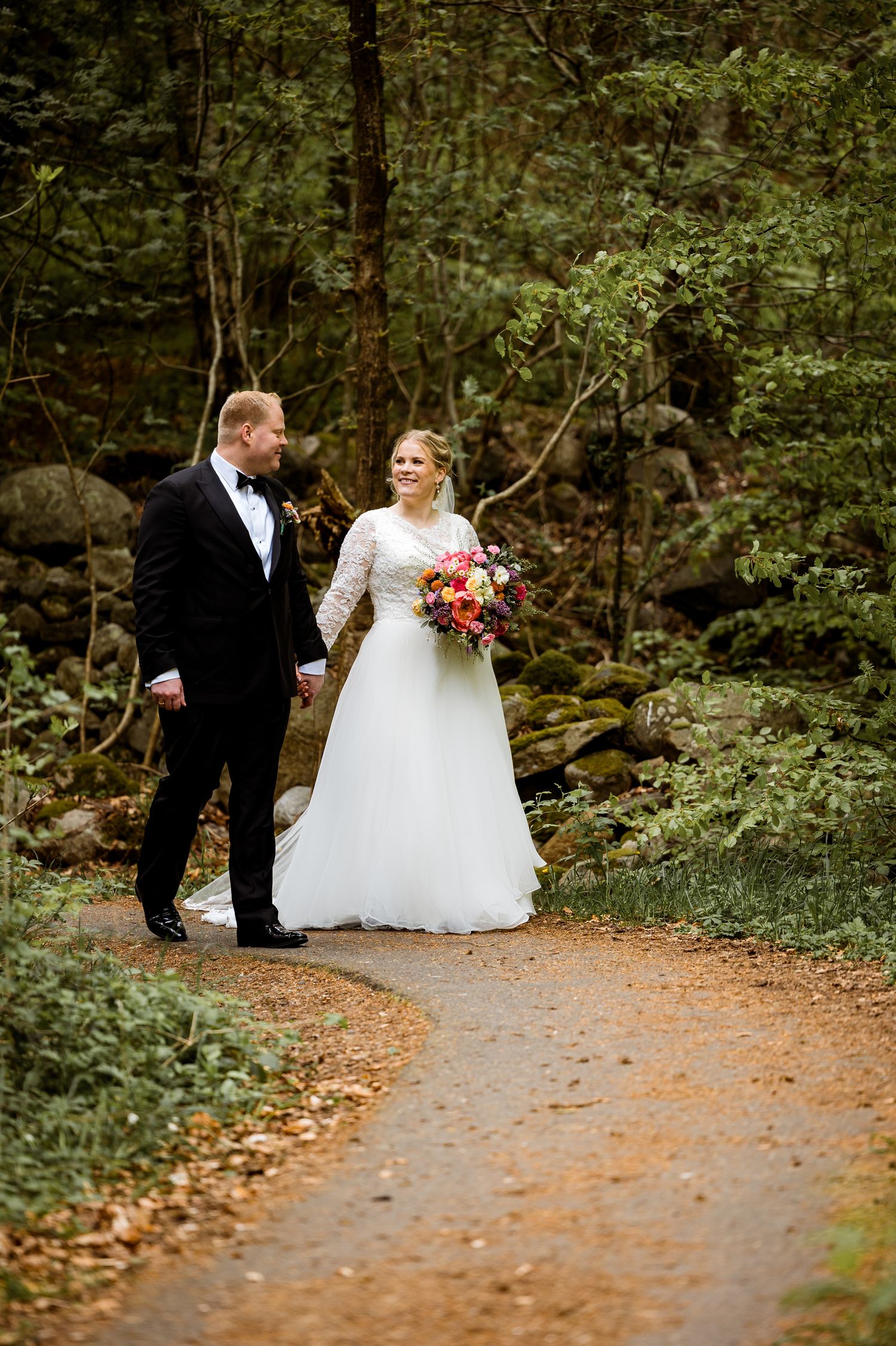 A romantic wedding couple walks along a wooded forest path surrounded by lush greenery and fallen autumn leaves.