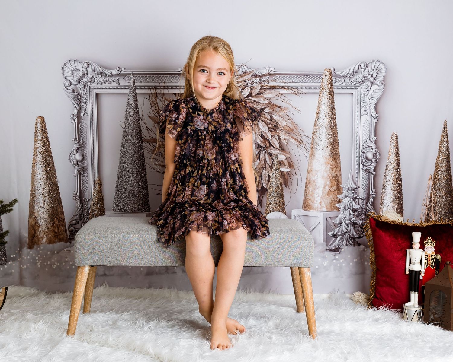 Child wearing black ruffled dress sits on gray bench with holiday backdrop.