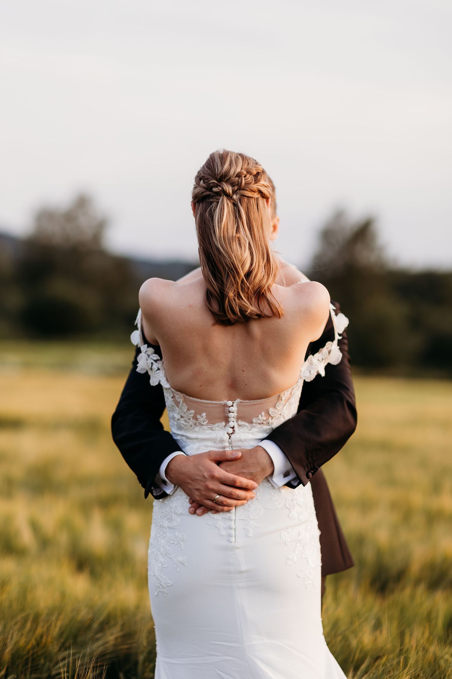 A romantic embrace from behind shows a bride in a white lace dress with braided hair standing in a golden field at sunset.
