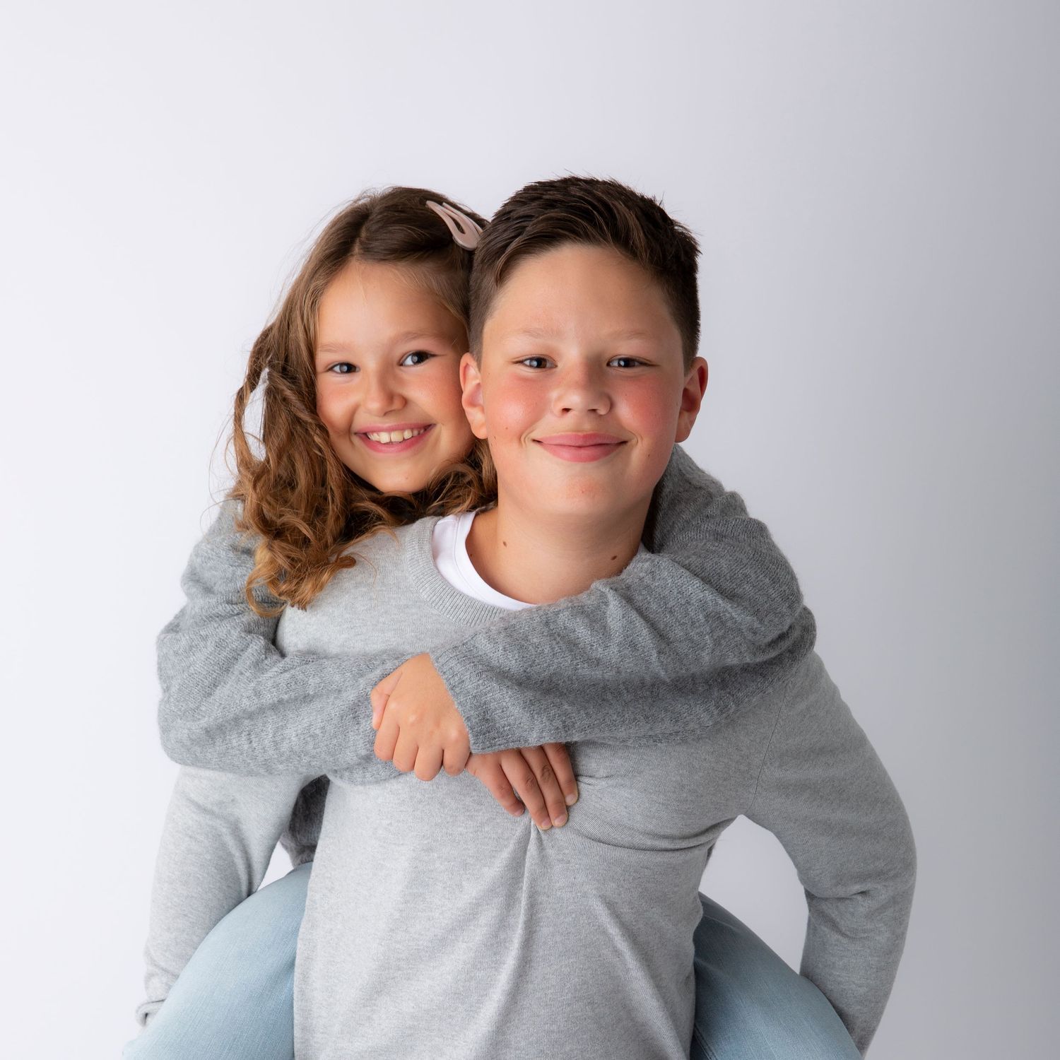 Siblings in matching gray sweatshirts hugging and smiling against white background.