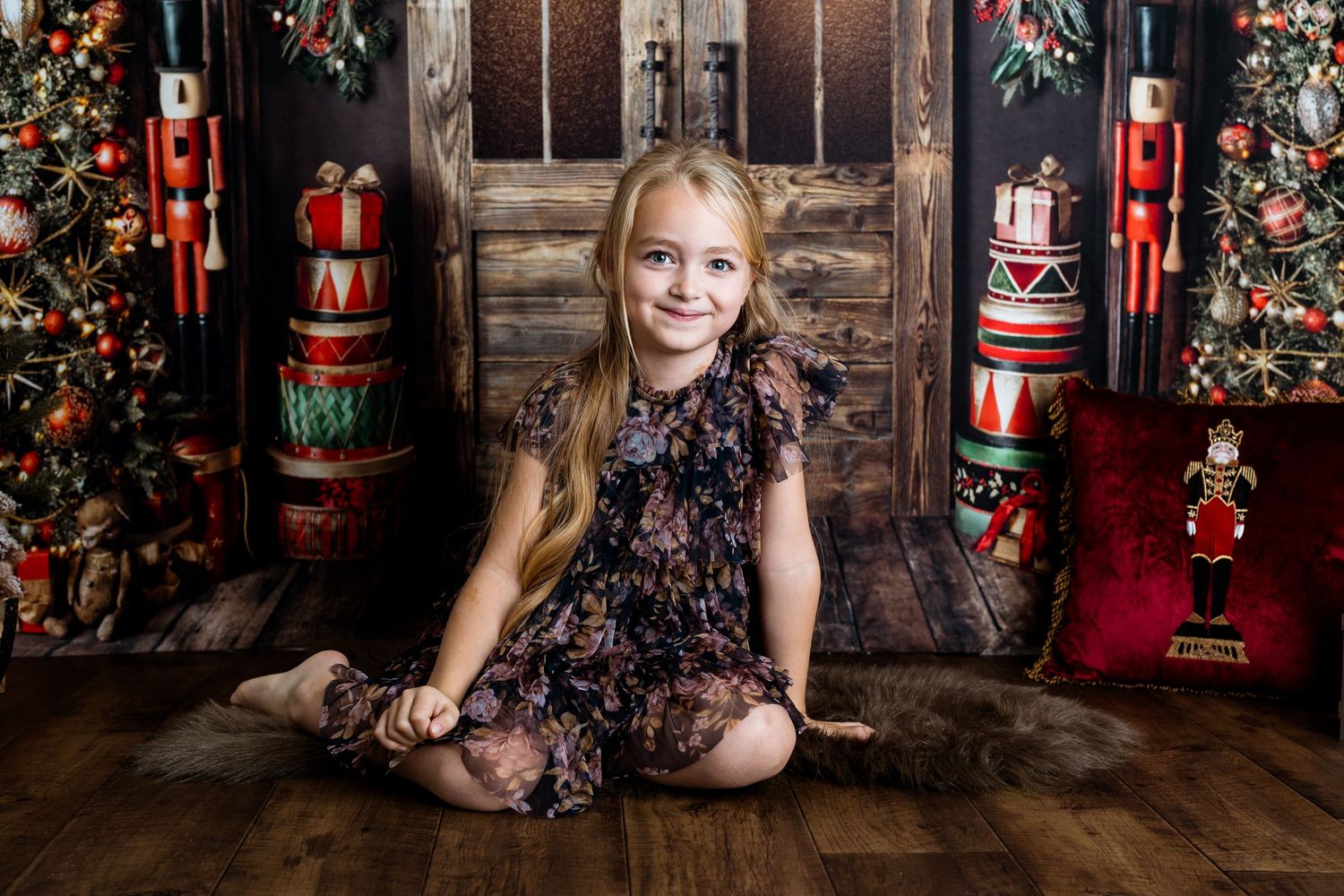 Child in dark floral dress sits on floor with vintage Christmas decorations and nutcrackers.