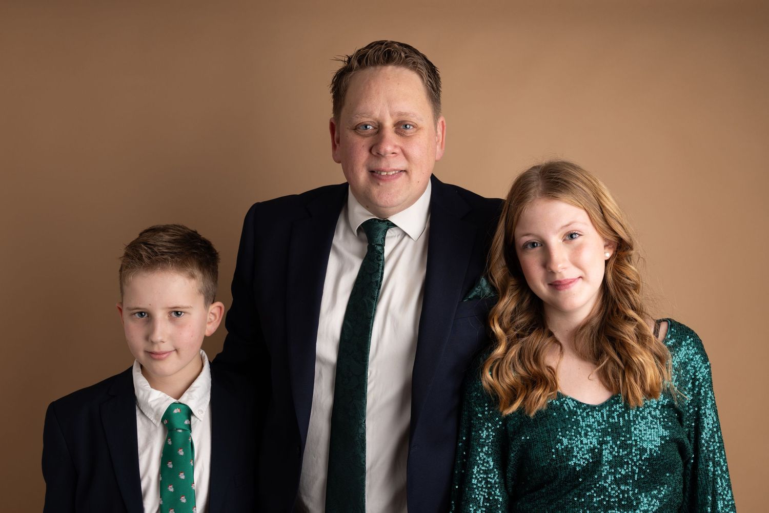 A formal portrait of three people in elegant attire against a brown backdrop, featuring emerald green and black clothing.