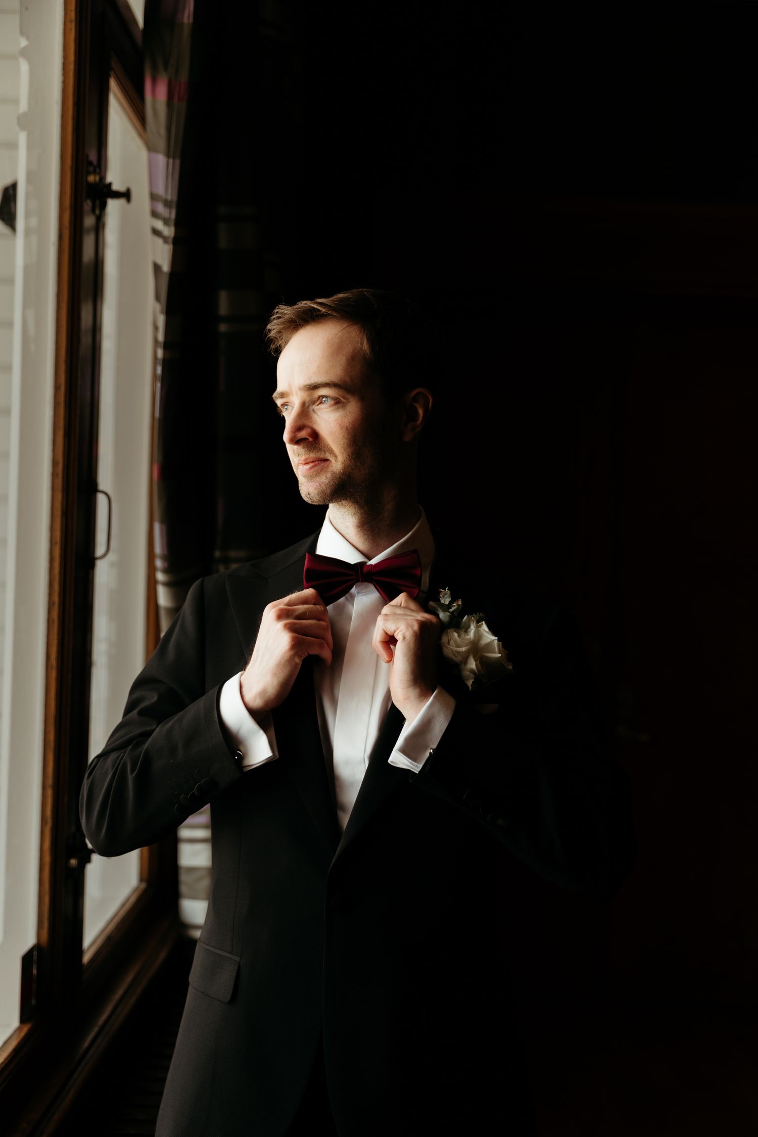 A moody portrait of groom adjusting bowtie by window light in classic tuxedo.