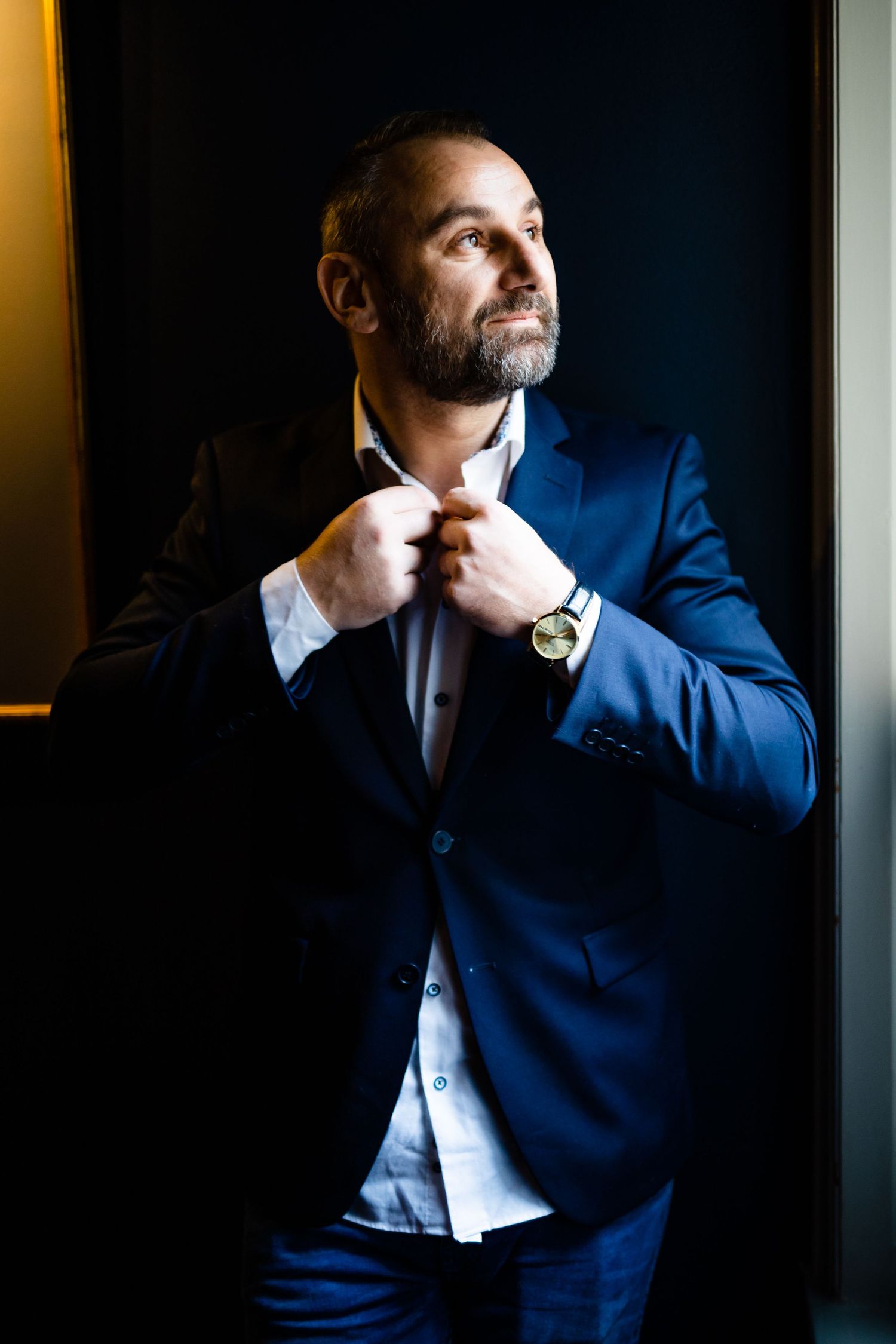 Groom in navy suit poses thoughtfully by window in dramatic lighting.