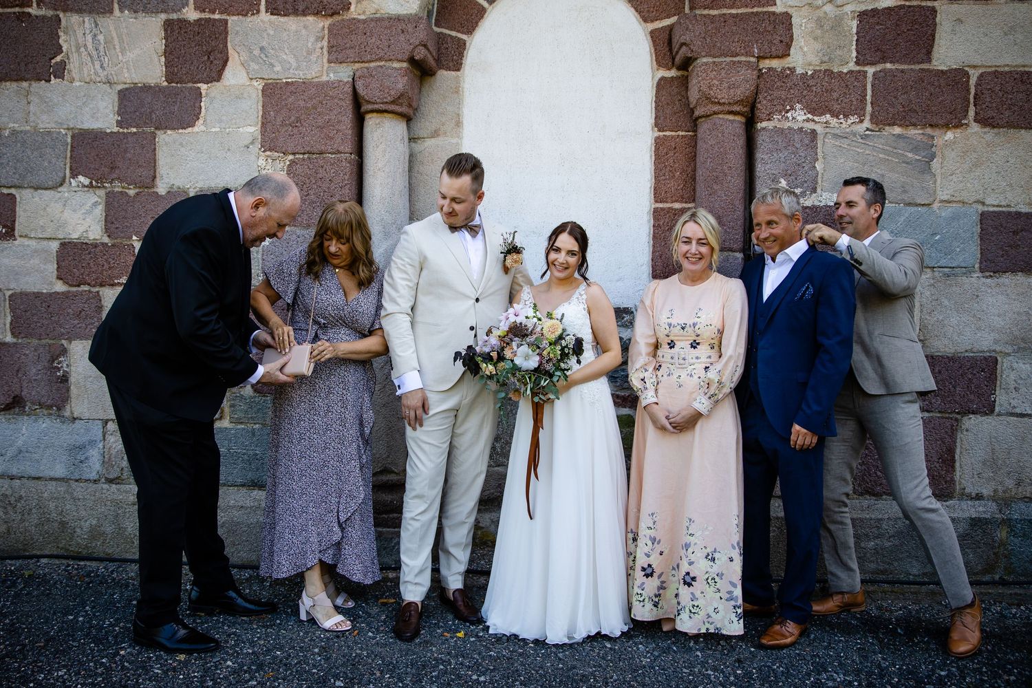 Wedding party poses casually against a rustic stone wall for a group photo.