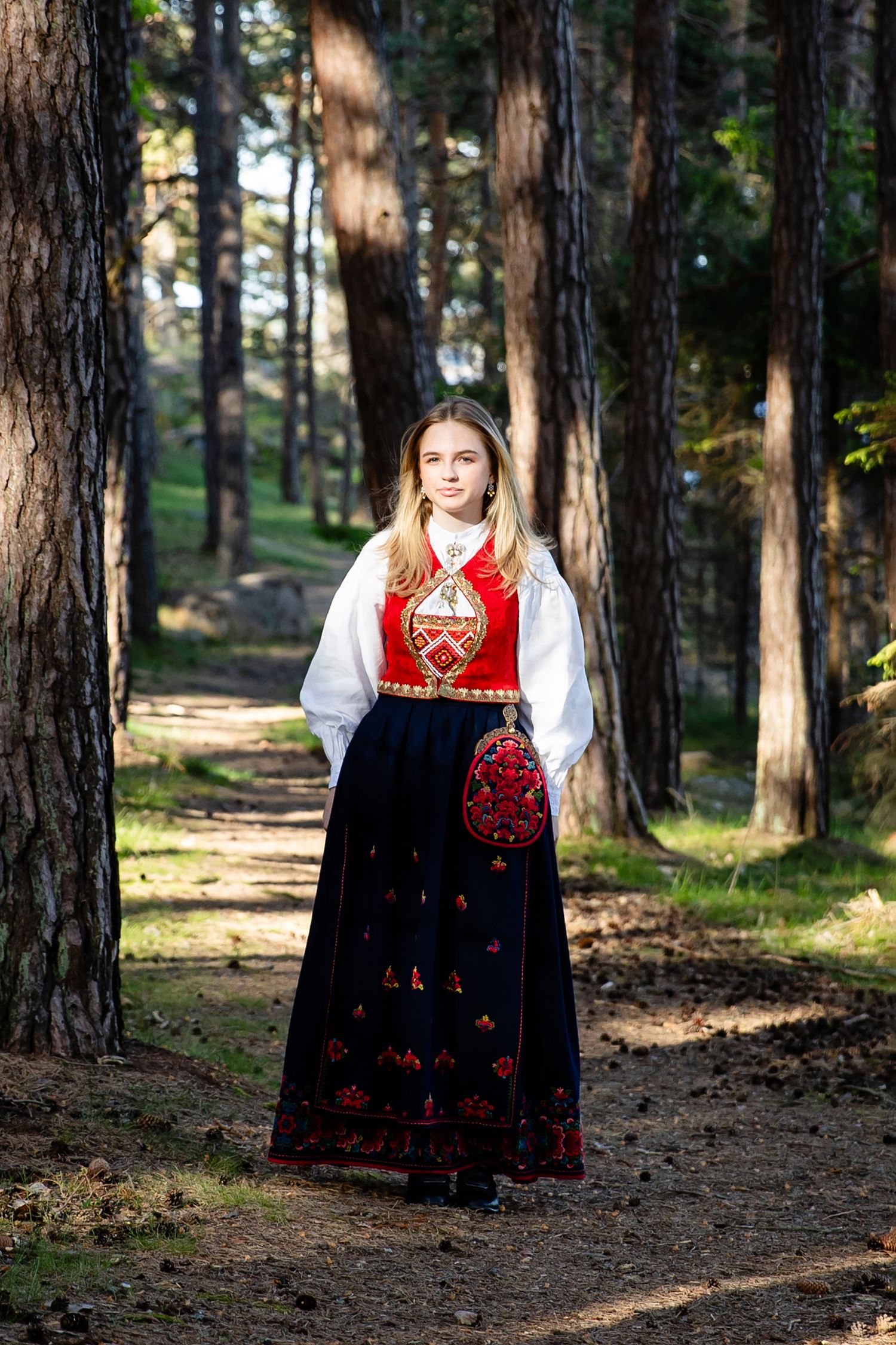 Traditional Norwegian bunad with red vest and black embroidered skirt shown on forest path.
