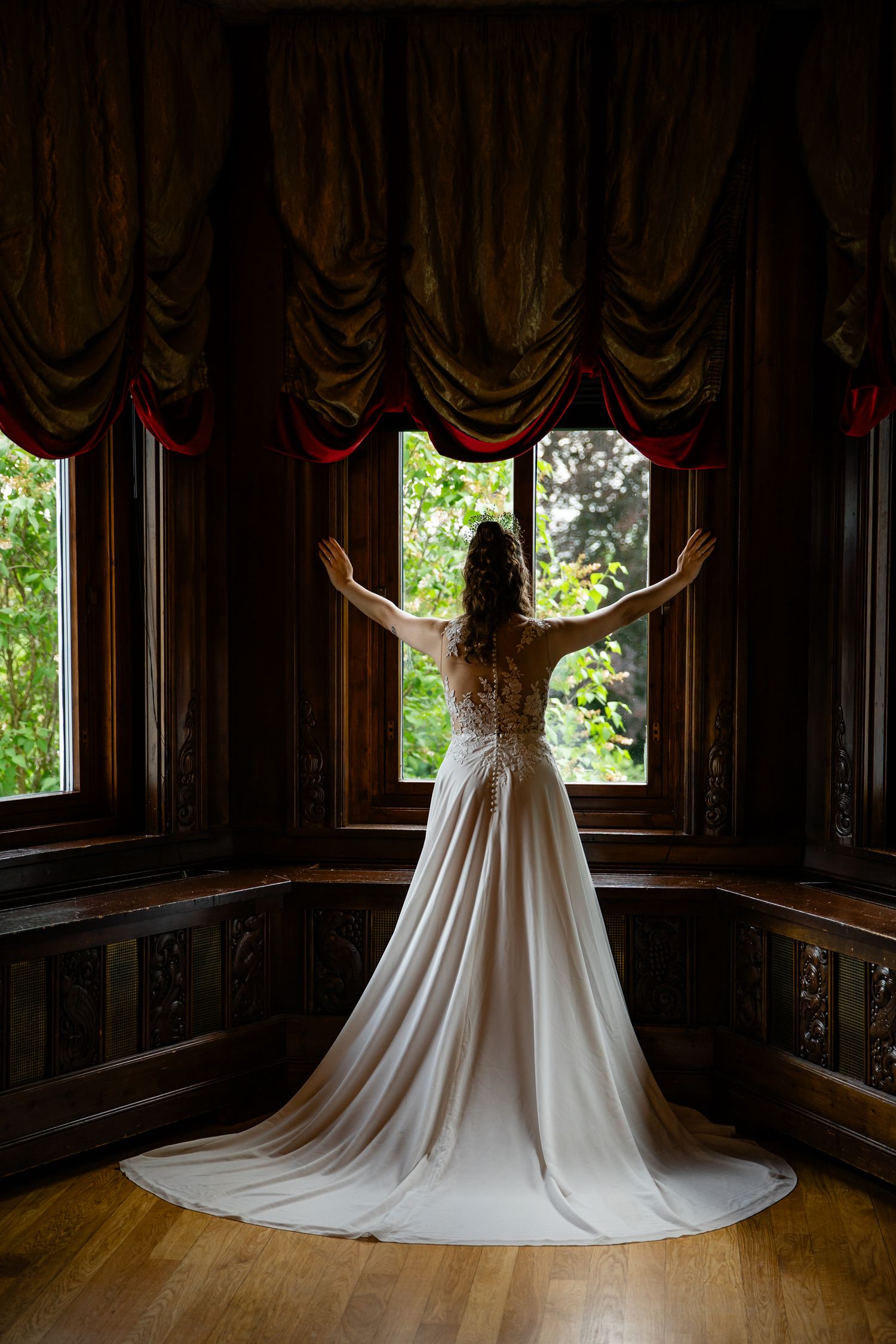 Wedding dress displayed in dramatic lighting against dark wood paneled room with windows.
