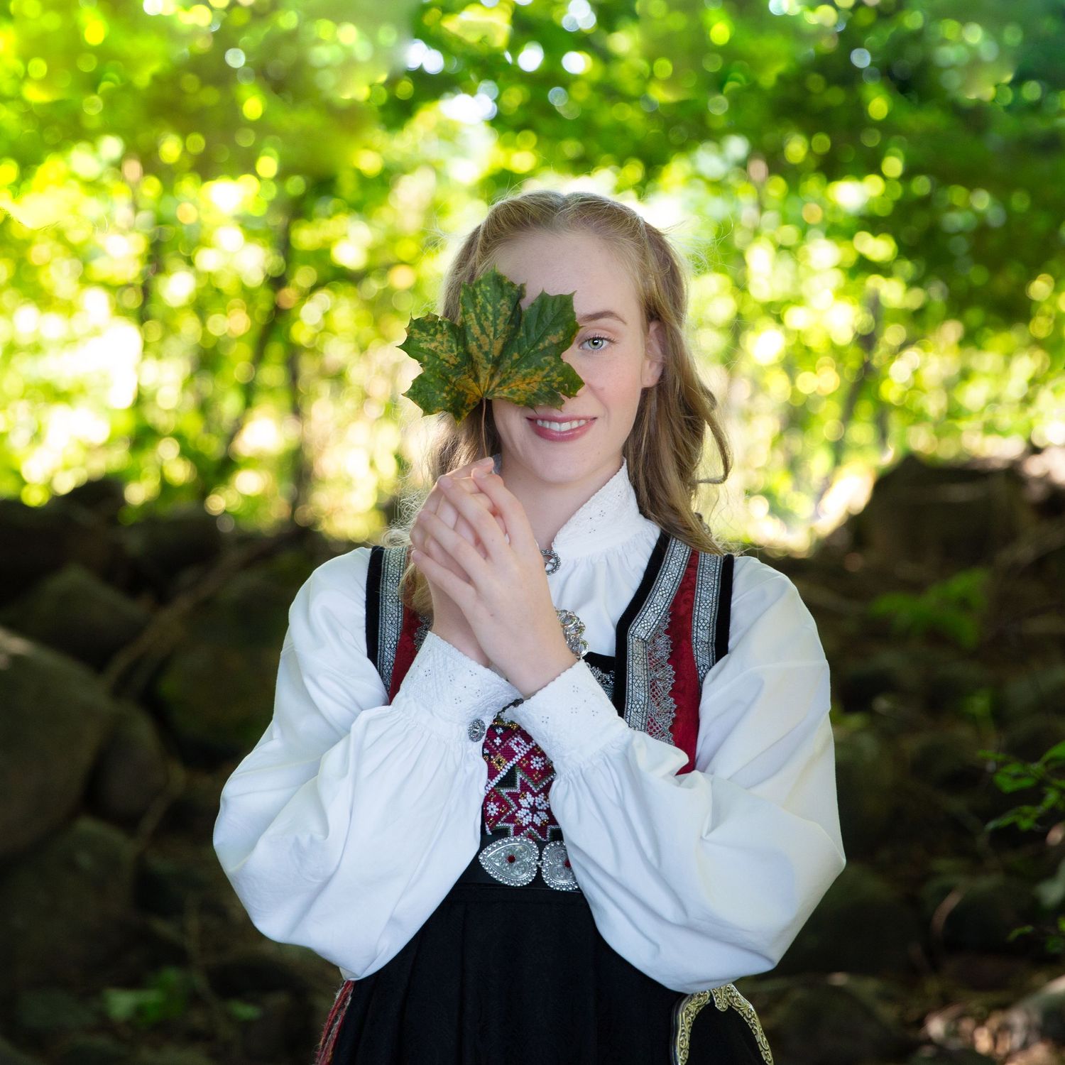 A person in traditional dress holds a large green leaf in front of their face while standing in a forest.