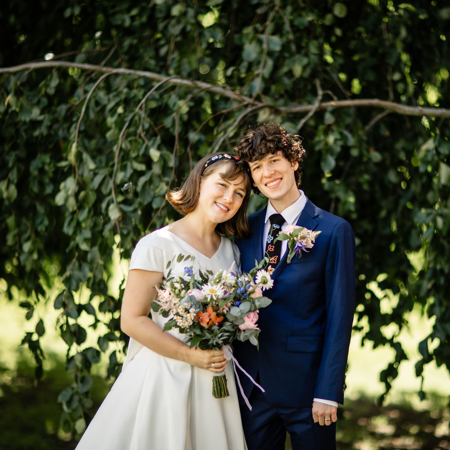 Wedding couple sharing joyful moment under trees with floral bouquet.