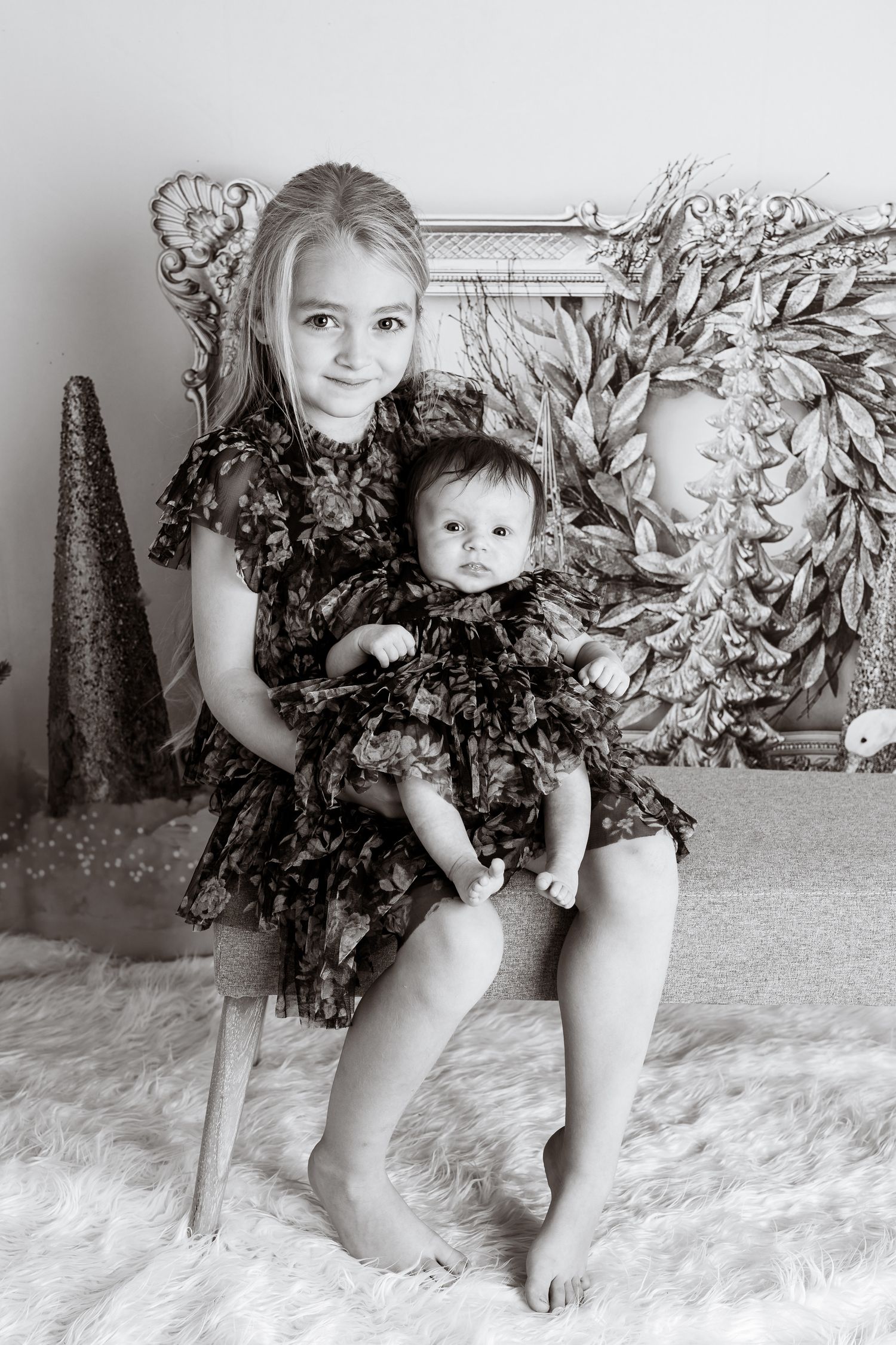 Two children wearing matching dark floral dresses pose together on bench with holiday decor.