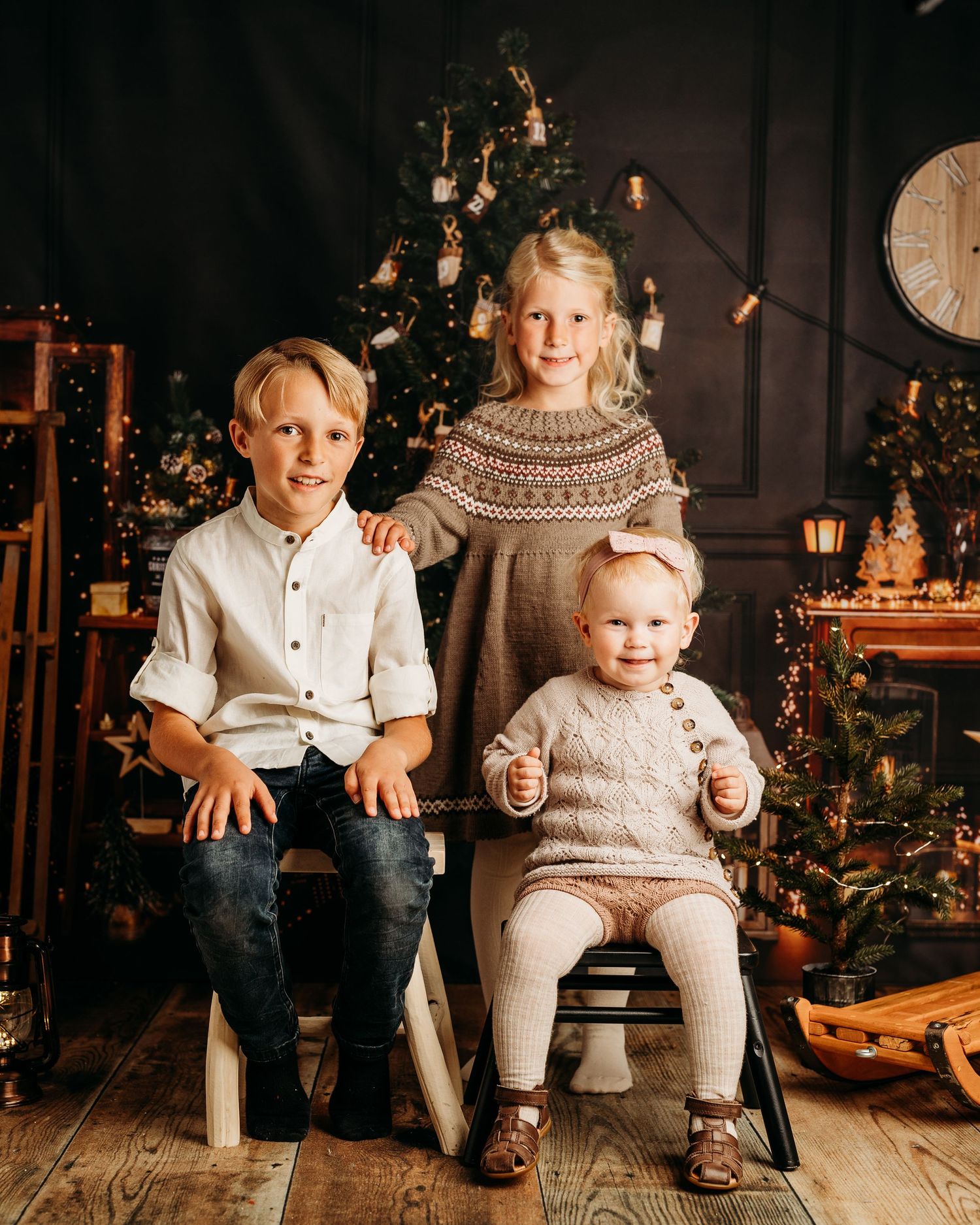 Playful holiday portraits with siblings sitting on wooden floor by Christmas tree.