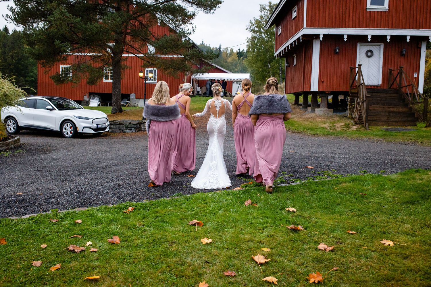 Wedding group gathers outside traditional red barn with autumn leaves scattered on ground.