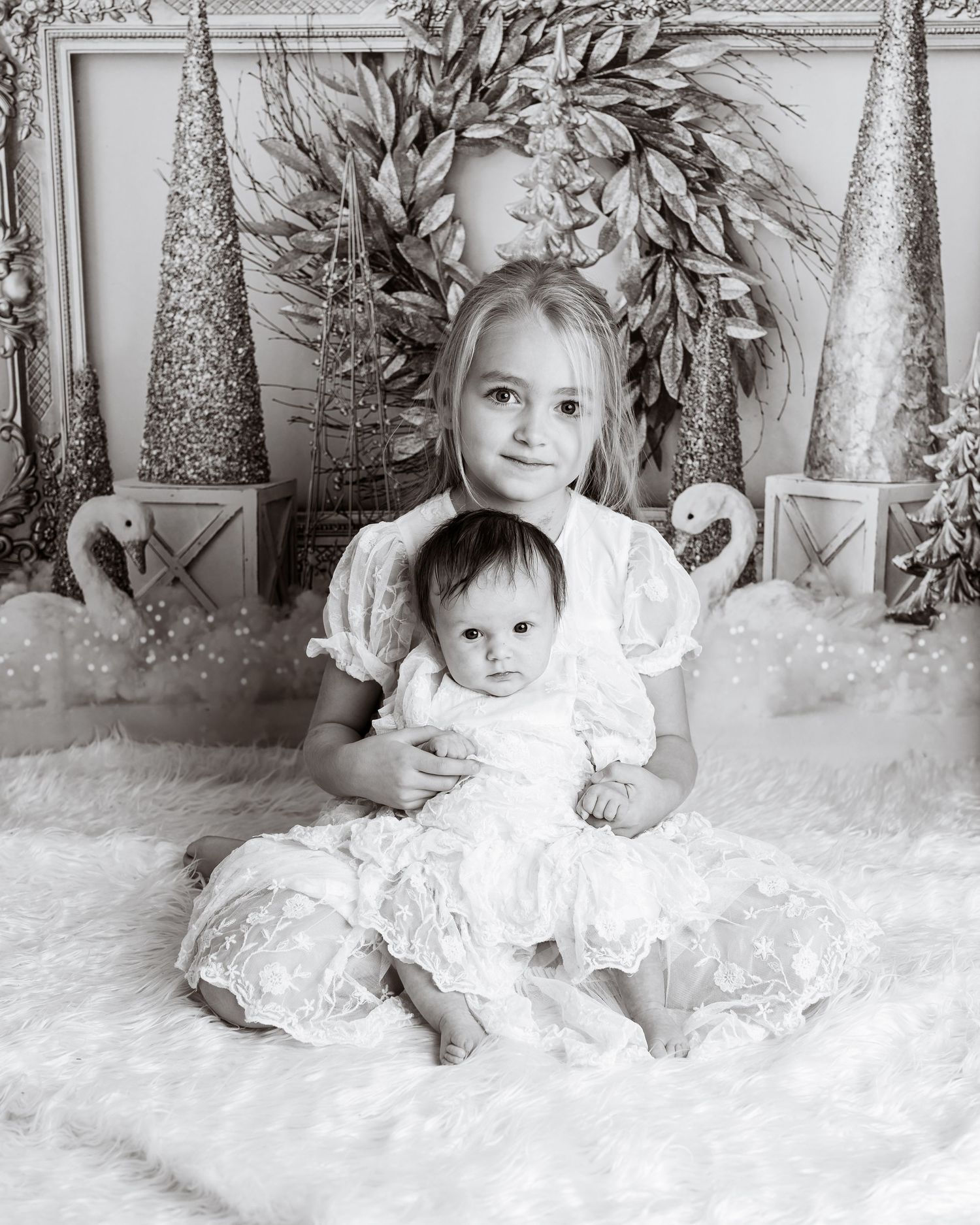 Black and white holiday photo of two children in matching white dresses with festive decor.