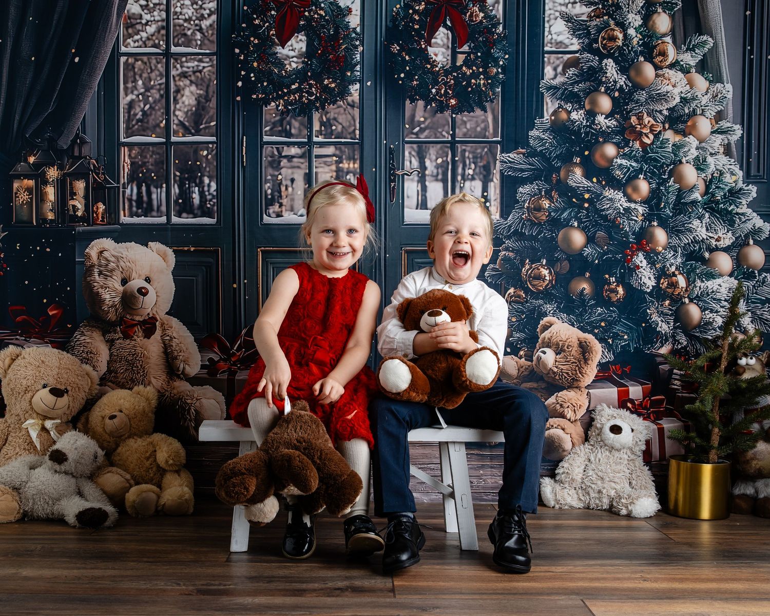 Two joyful children sit on a bench surrounded by teddy bears near a decorated Christmas tree in a festive holiday portrait.