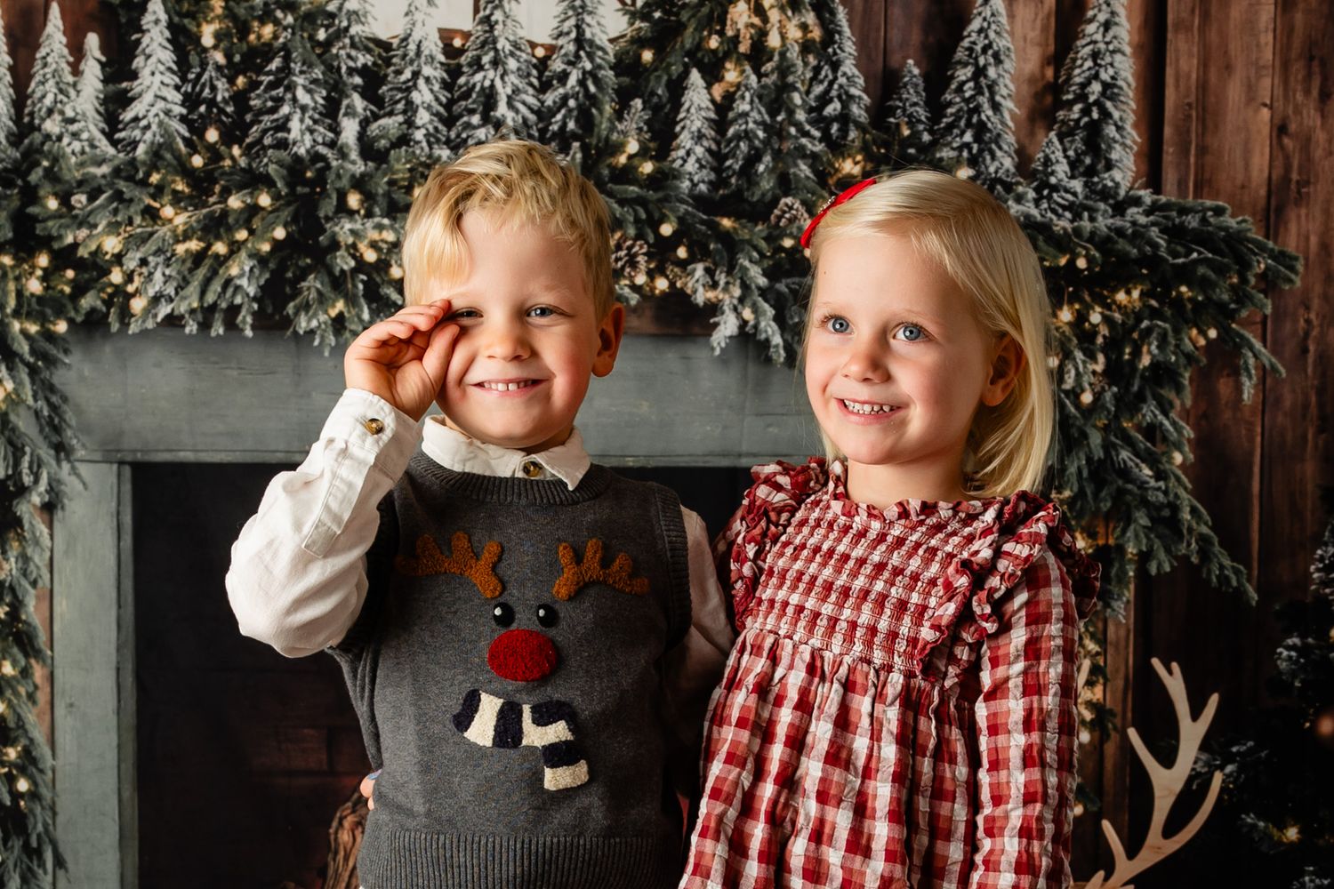 Two smiling children in a reindeer sweater and a red plaid dress pose by a decorated fireplace with a Christmas garland.