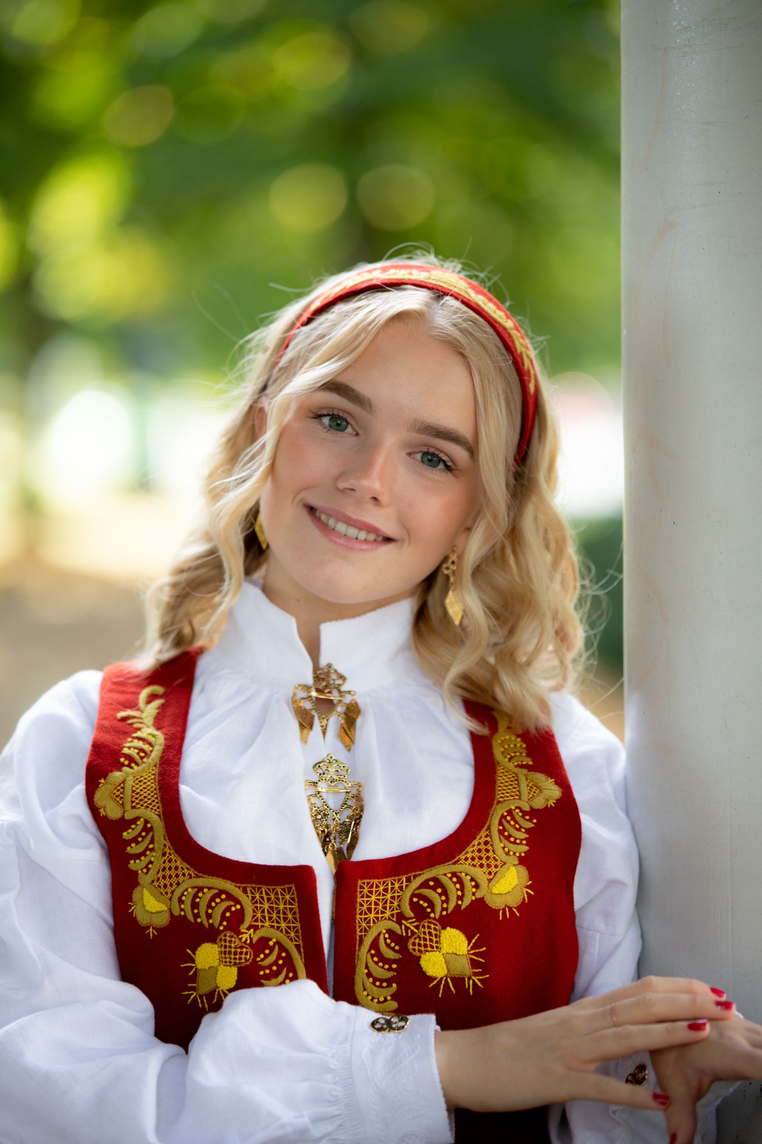 Traditional Norwegian bunad dress with red and gold embroidered vest and white blouse shown in outdoor portrait.