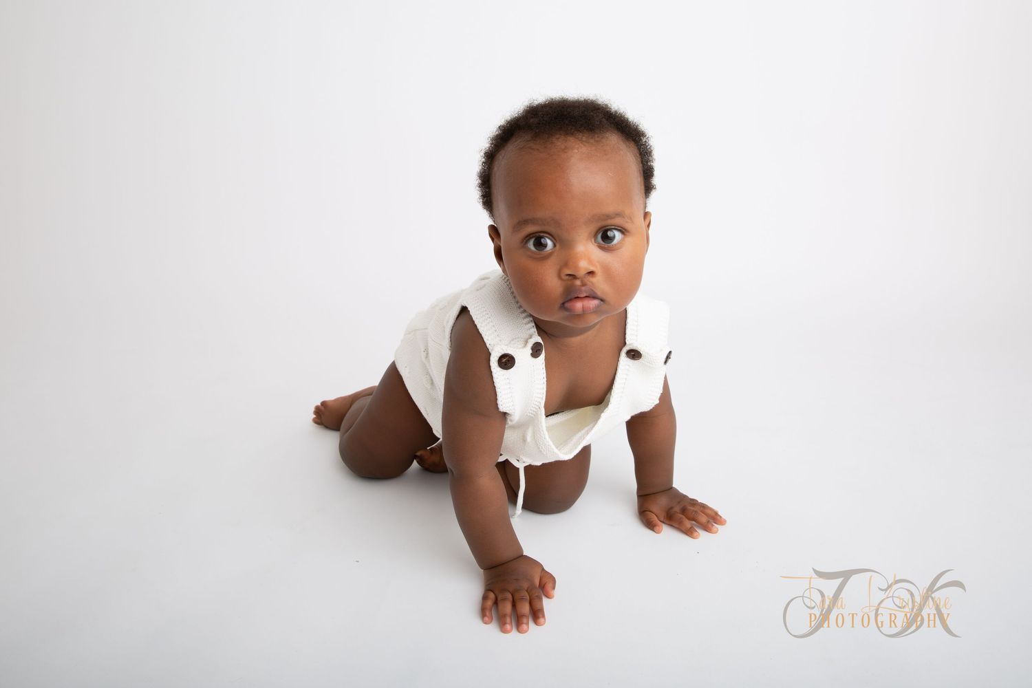 Babyfotografering Moss: En baby krabber mot kameraet på hvit bakgrunn, kledd i en hvit strikket drakt.