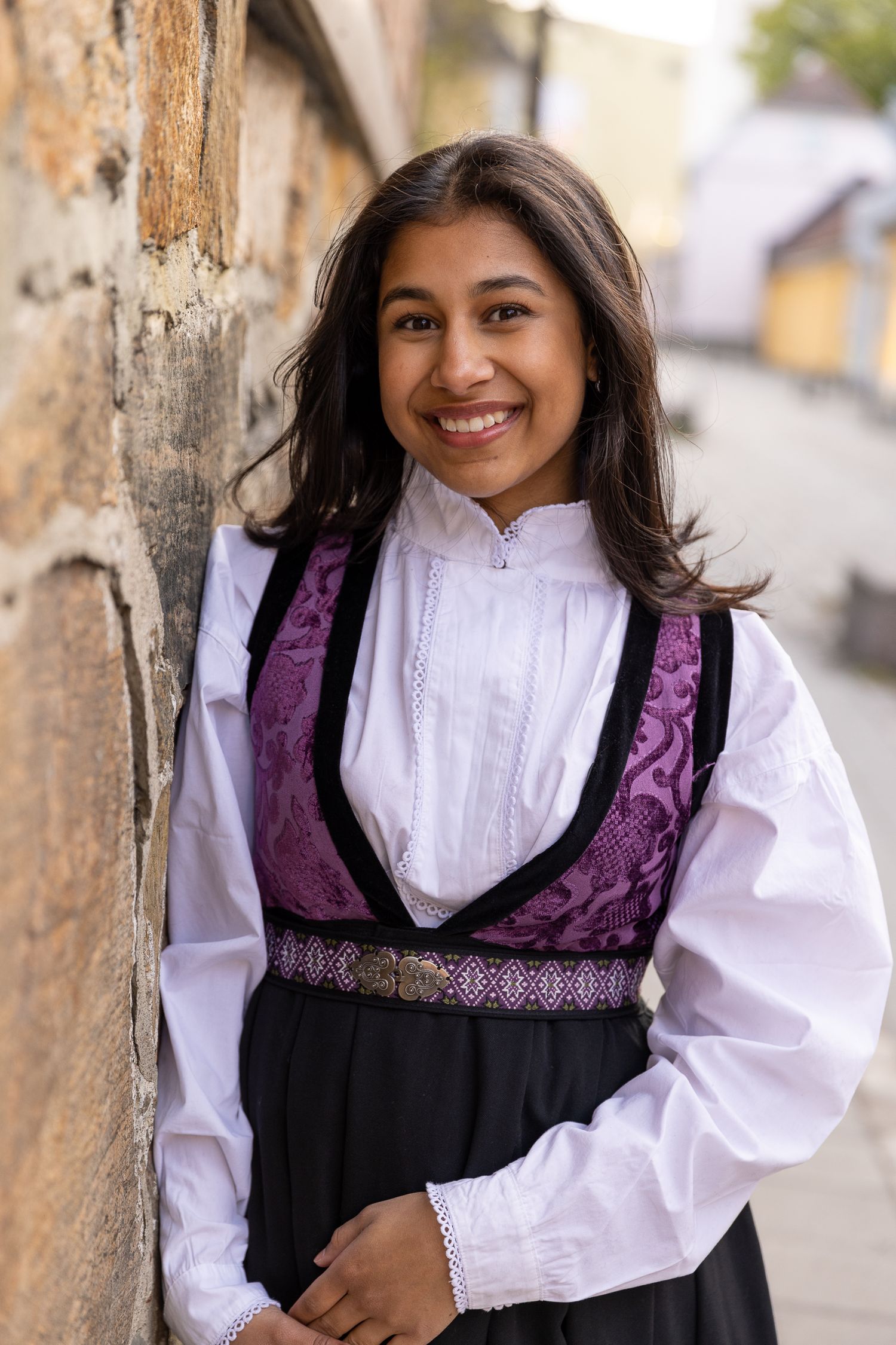 Portrait of someone wearing a traditional black and purple embroidered dress with white blouse against a textured wall.