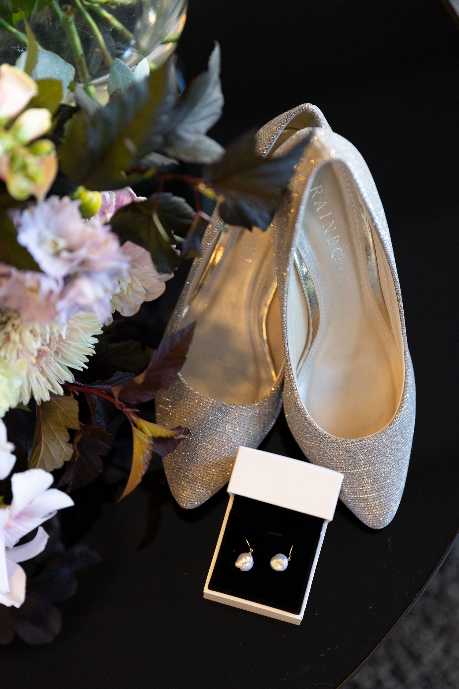 Wedding accessories including sparkly flat shoes, pearl earrings and delicate flowers arranged on black surface.