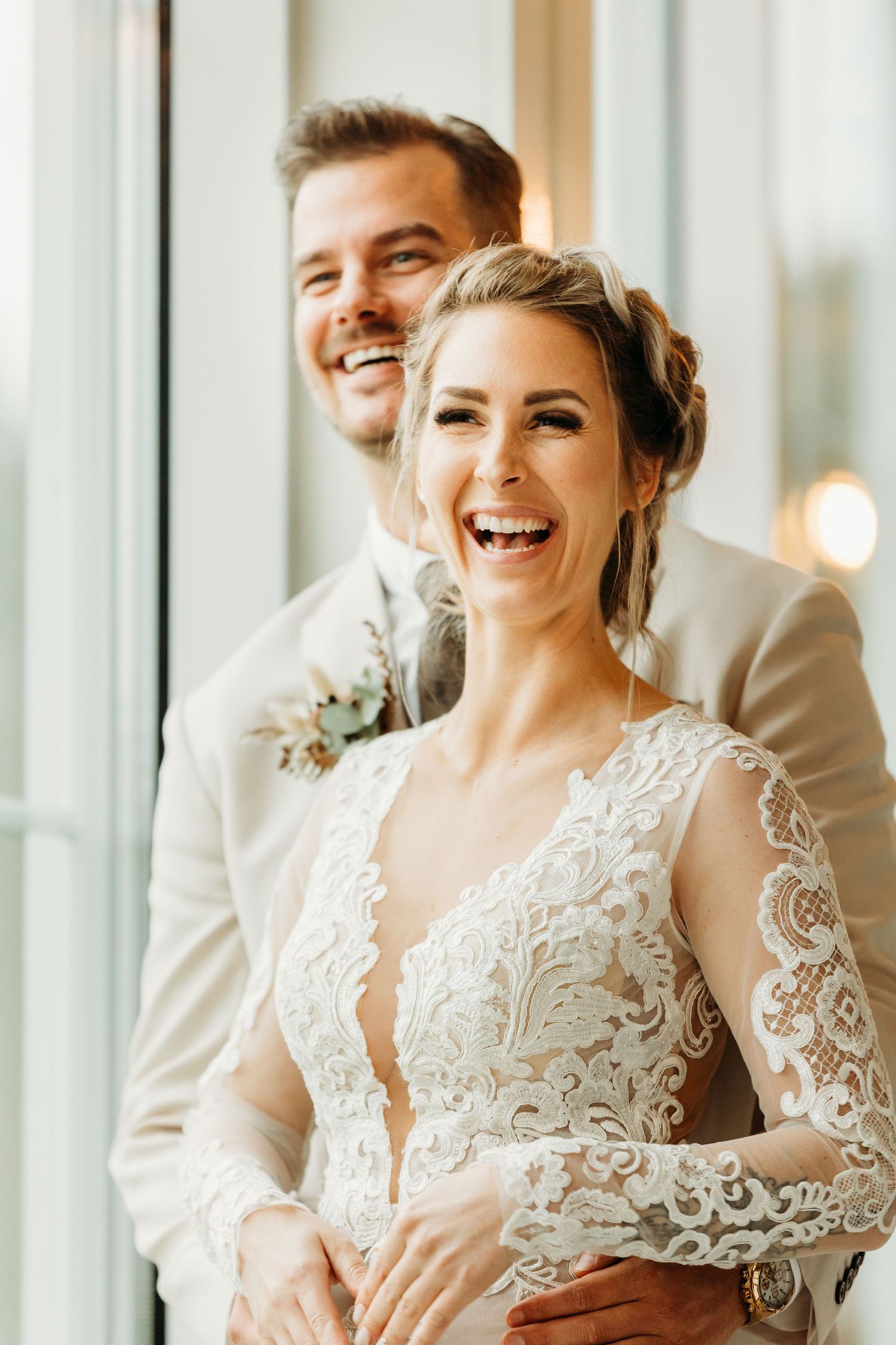 Joyful couple sharing laughter in front of bright windows wearing elegant wedding attire.