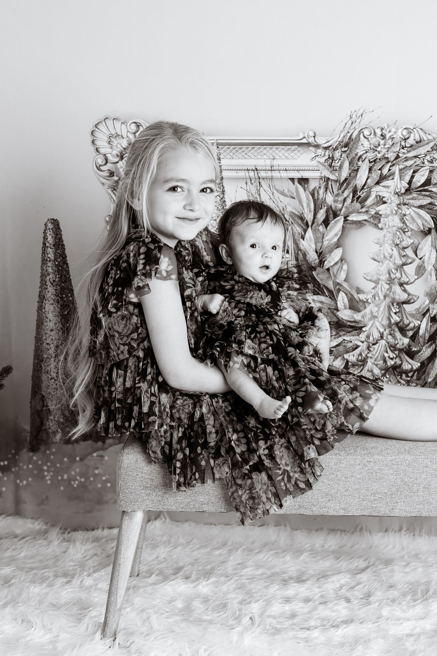 Black and white photo of two children in matching dark dresses against festive backdrop.
