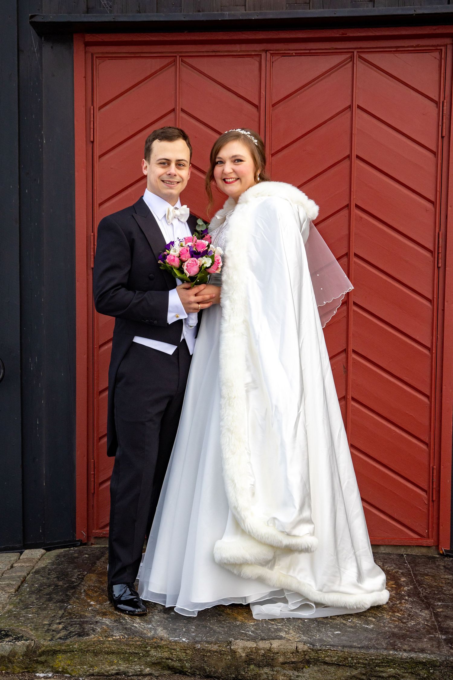 Two men in formal tuxedos pose back-to-back against stone wall at wedding.