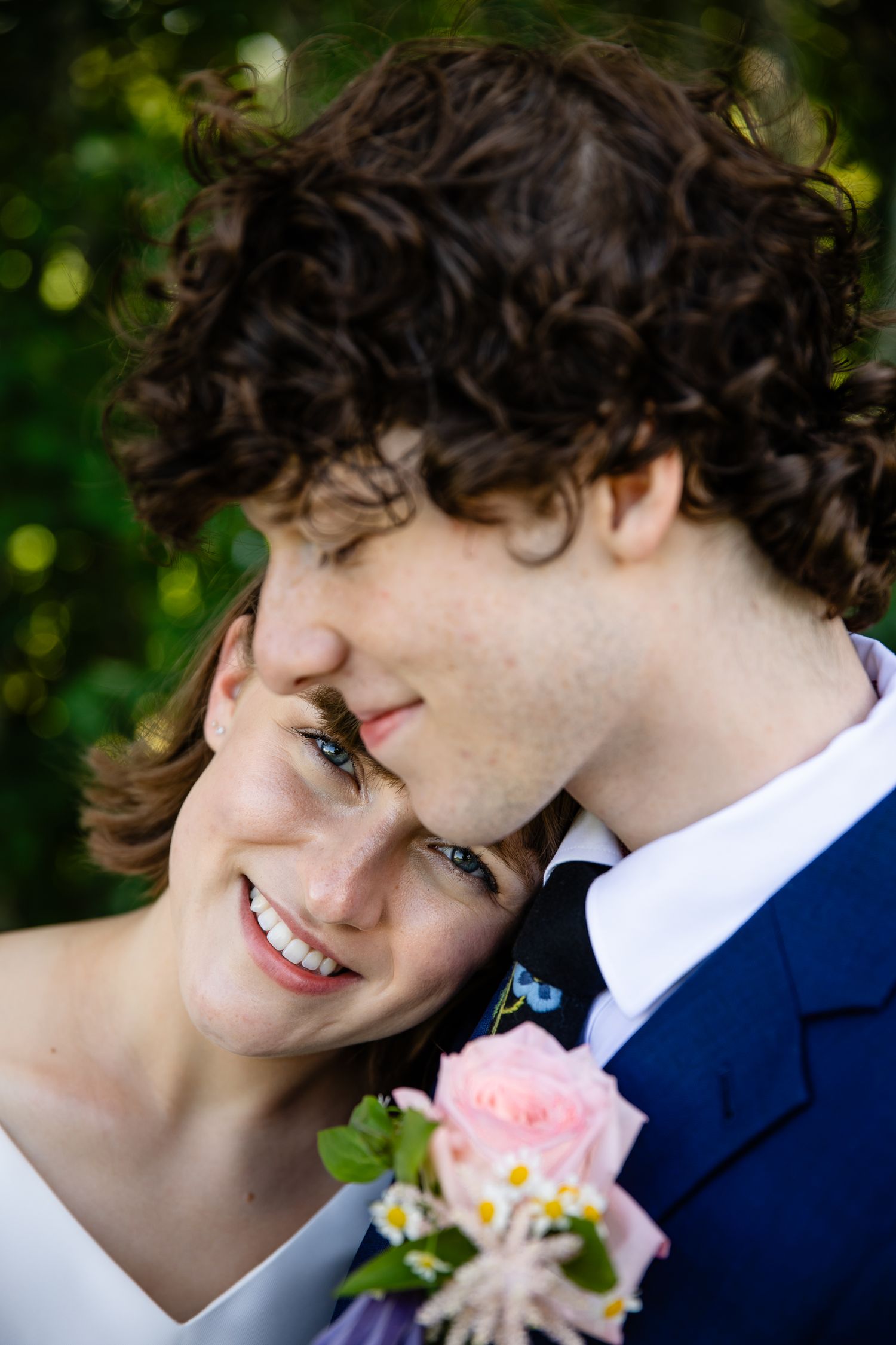 A romantic wedding portrait showing a tender moment with a pink rose boutonniere against a blurred green background.