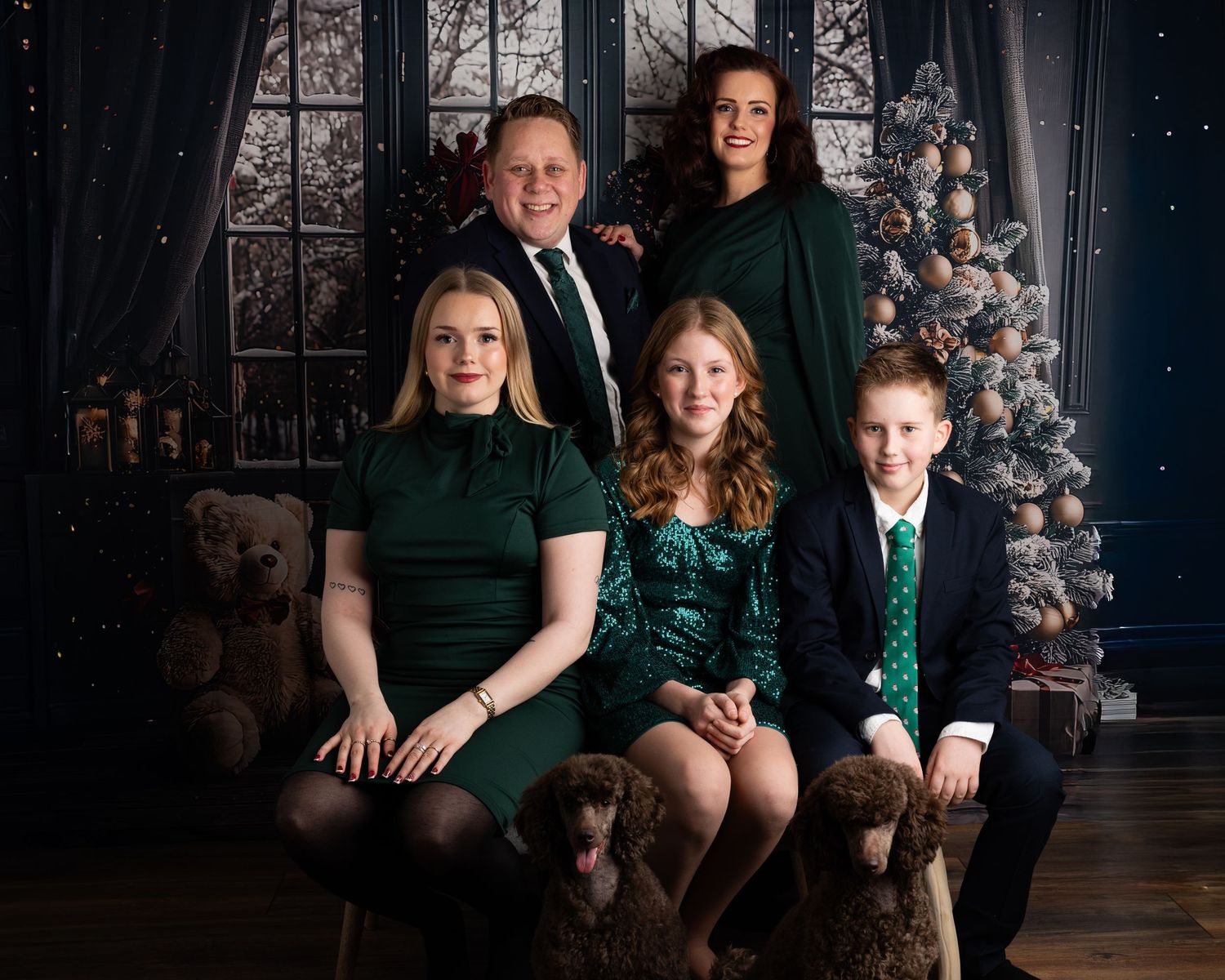 Family wearing harmonising green outfits poses together in a cozy rustic holiday setting with a winter window backdrop.