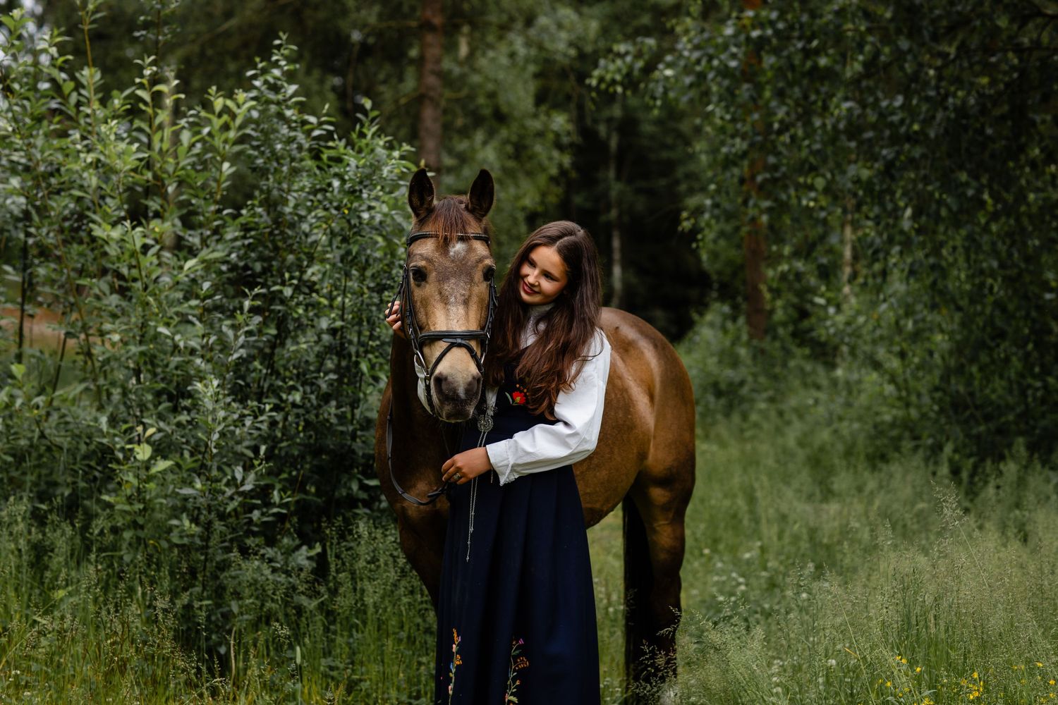 A brown horse and person in a white top and navy skirt stand together in a lush green forest setting.