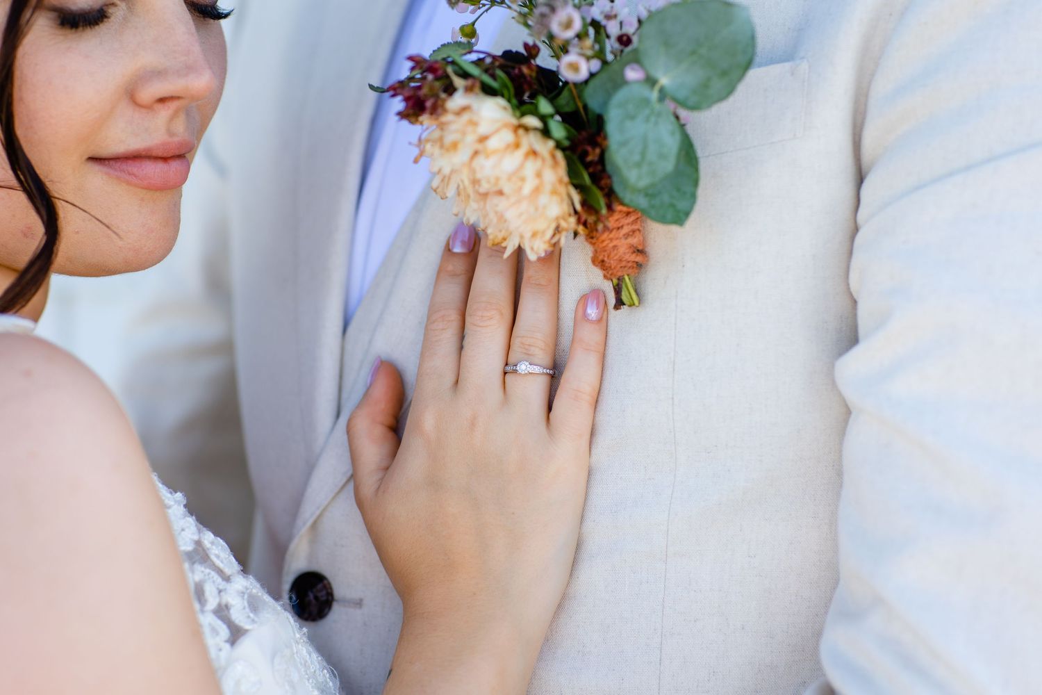Close up of a bride's hand with wedding ring resting on groom's white suit jacket with floral boutonniere.