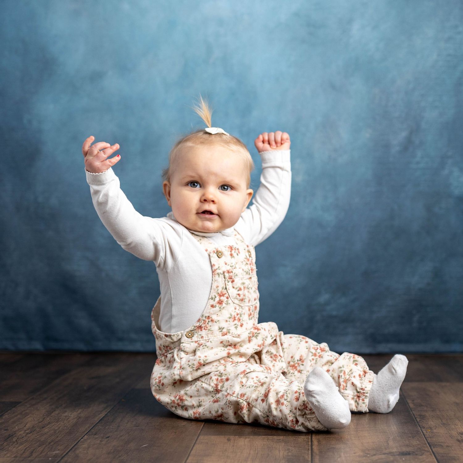 Babyfotograf Østfold: En begeistret baby i blomstrete snekkerbukse sitter på et mørkt tregulv med hendene i været mot en blå bakgrunn.