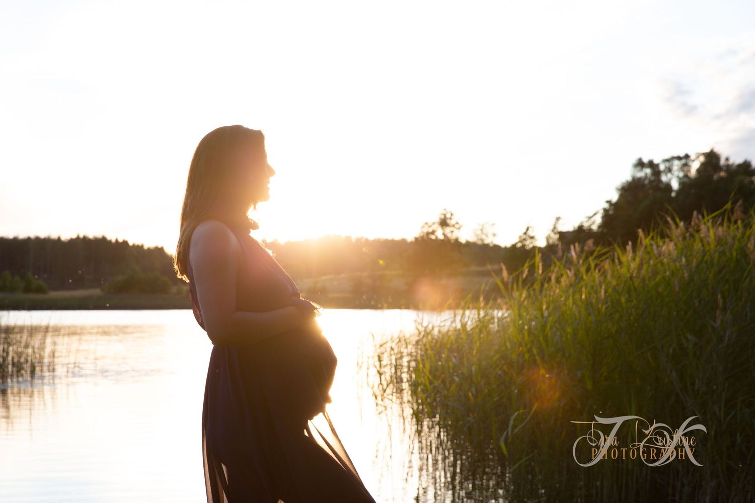 Silhouette of pregnant woman by lake at sunset with sun flare.