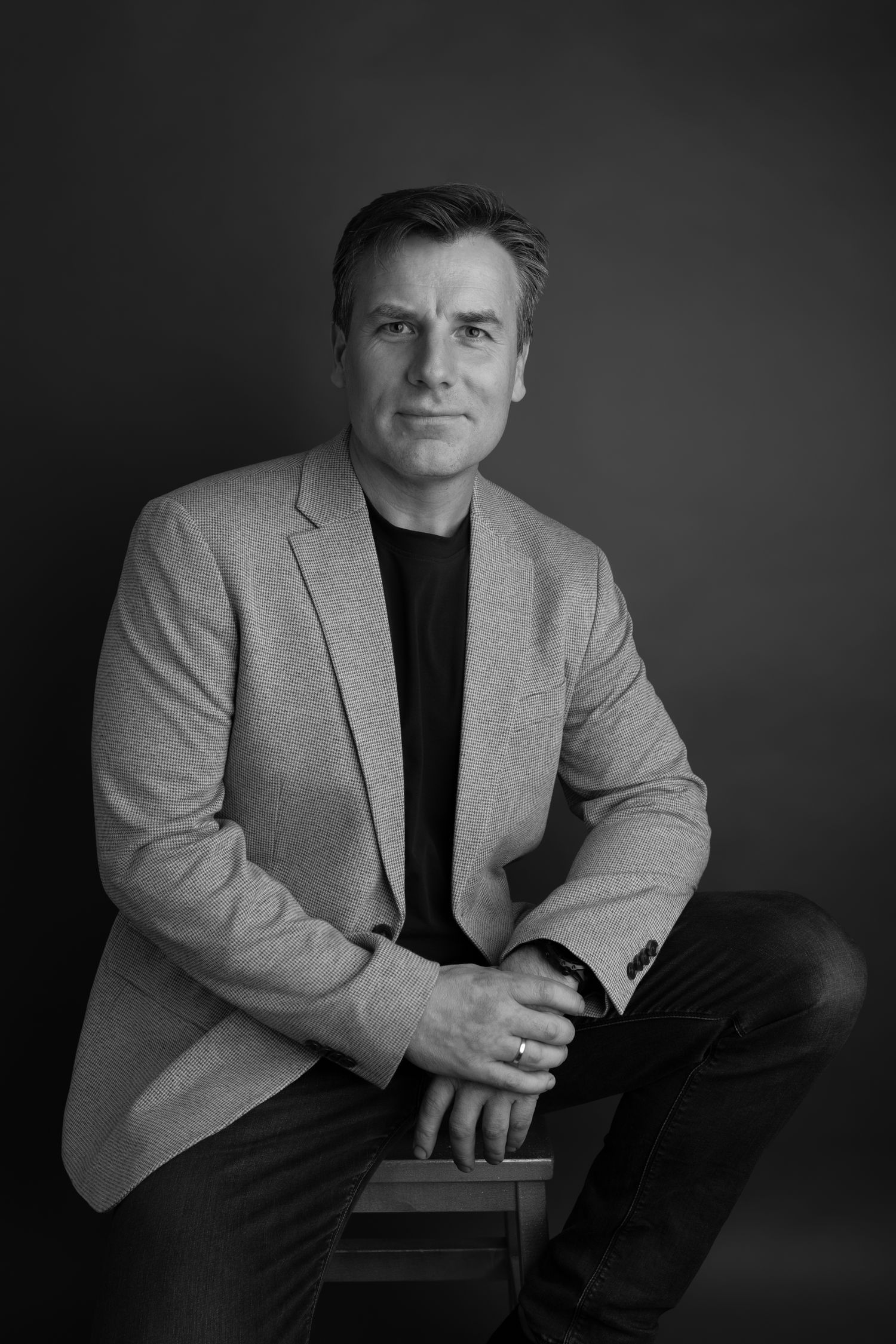 Professional black and white portrait of person in light blazer seated against dark background.