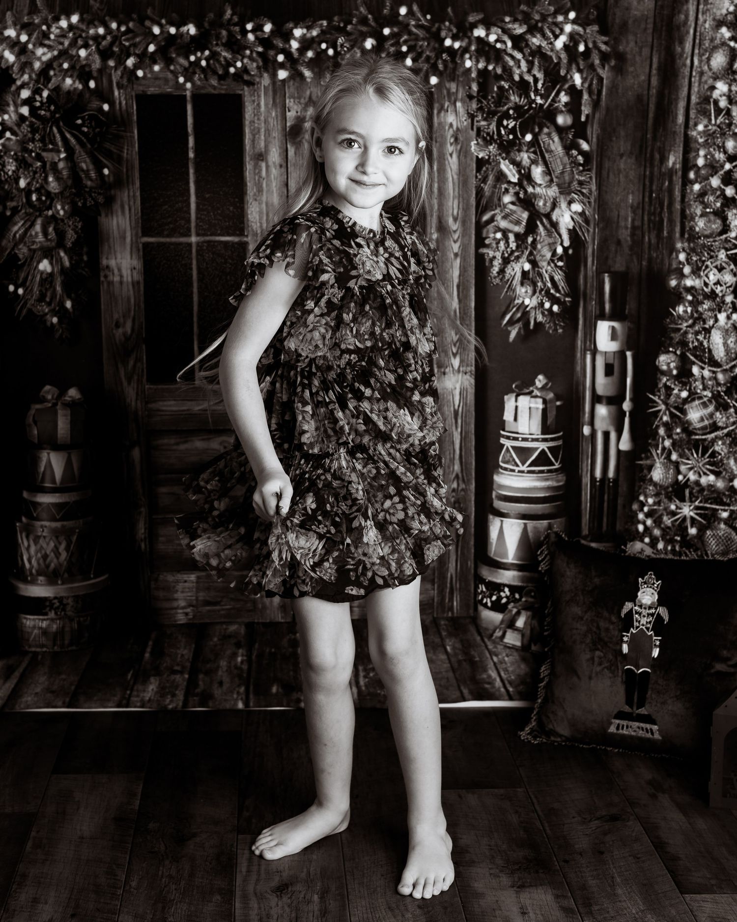 Black and white studio portrait against rustic wooden wall with decorative barrels and greenery.