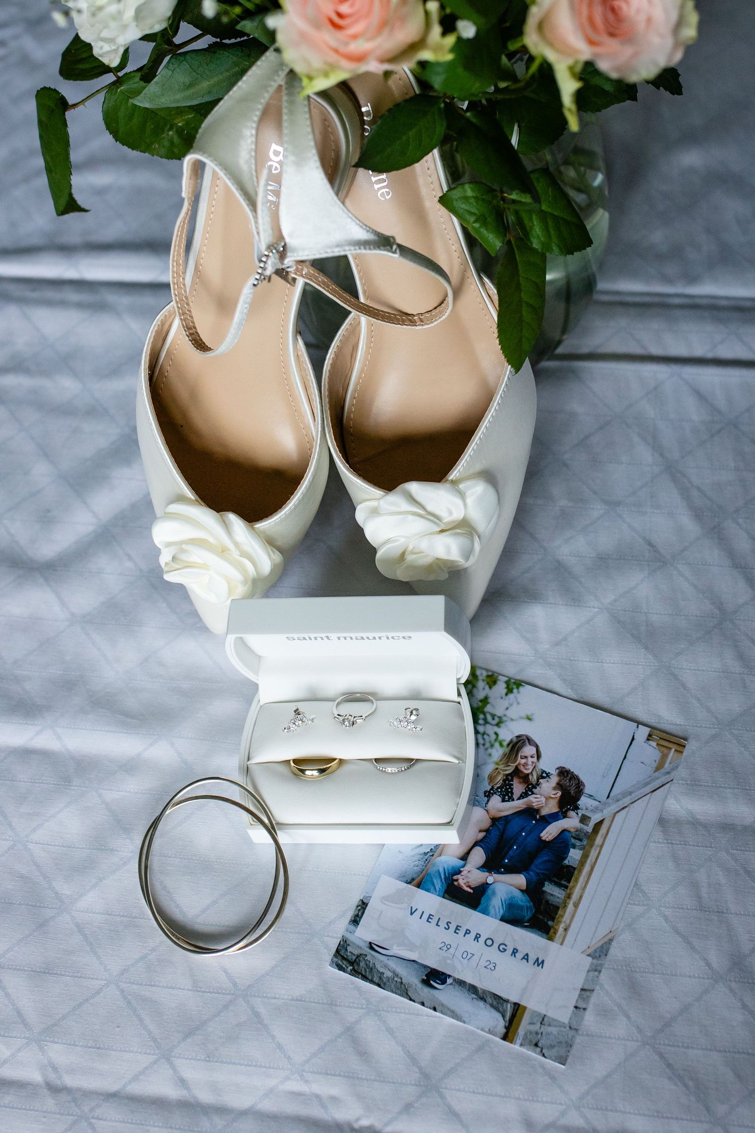 Wedding details with bridal shoes, roses, and engagement photos on gray fabric.