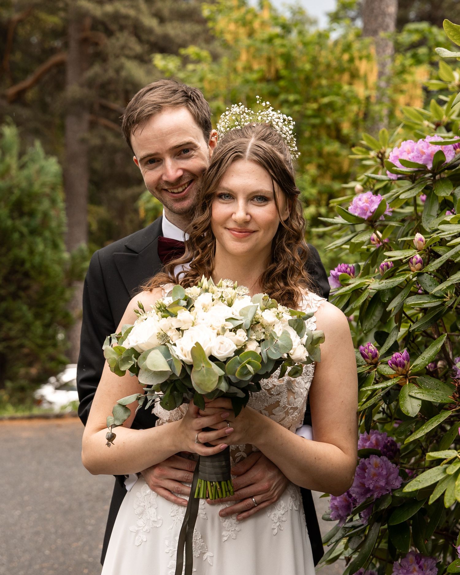 Newlyweds holding white floral bouquet together in a garden setting with purple flowers.