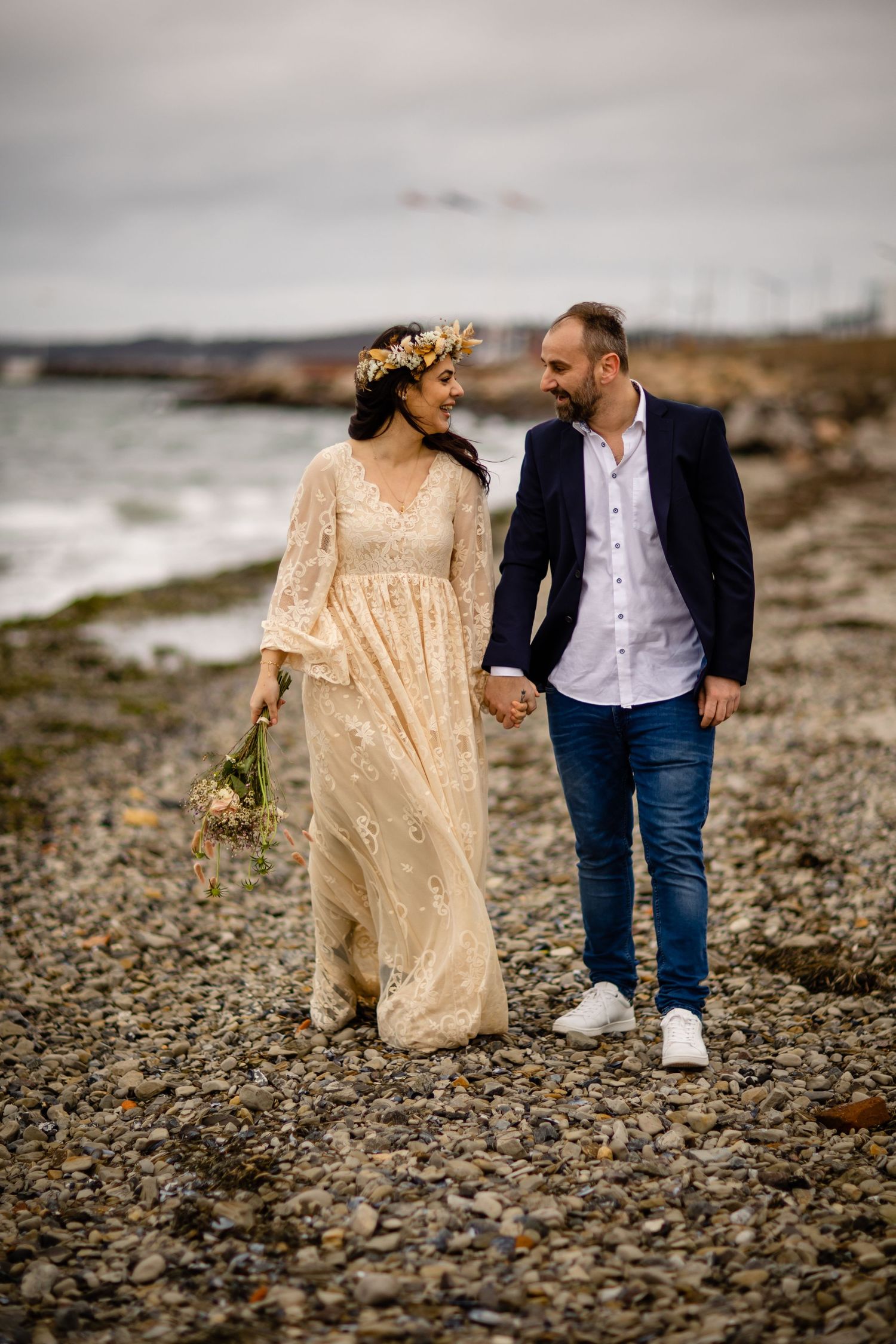 Couple walking along rocky beach shore in elegant wedding attire.