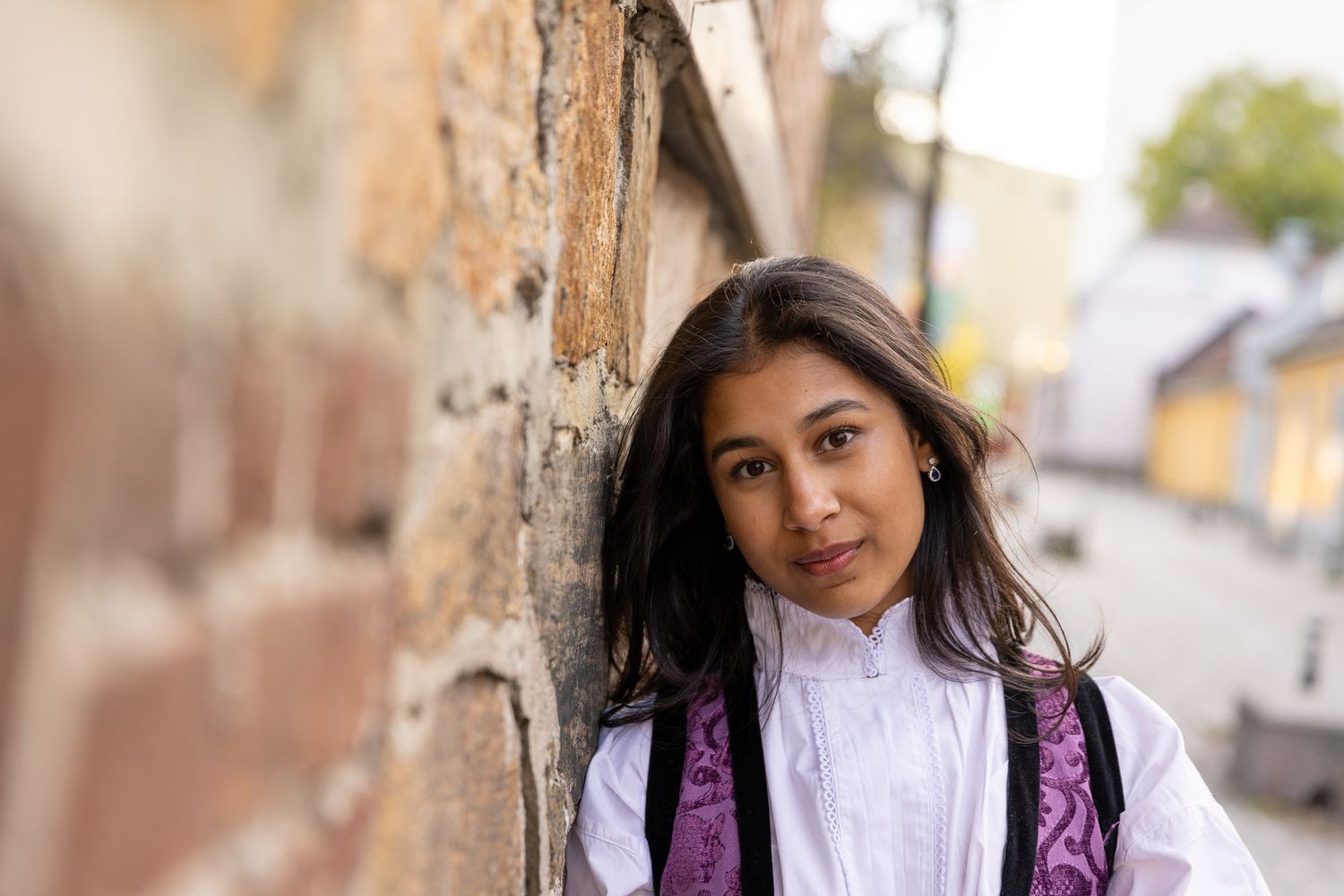 Person in traditional dress smiling warmly while posing against a rustic wall background.