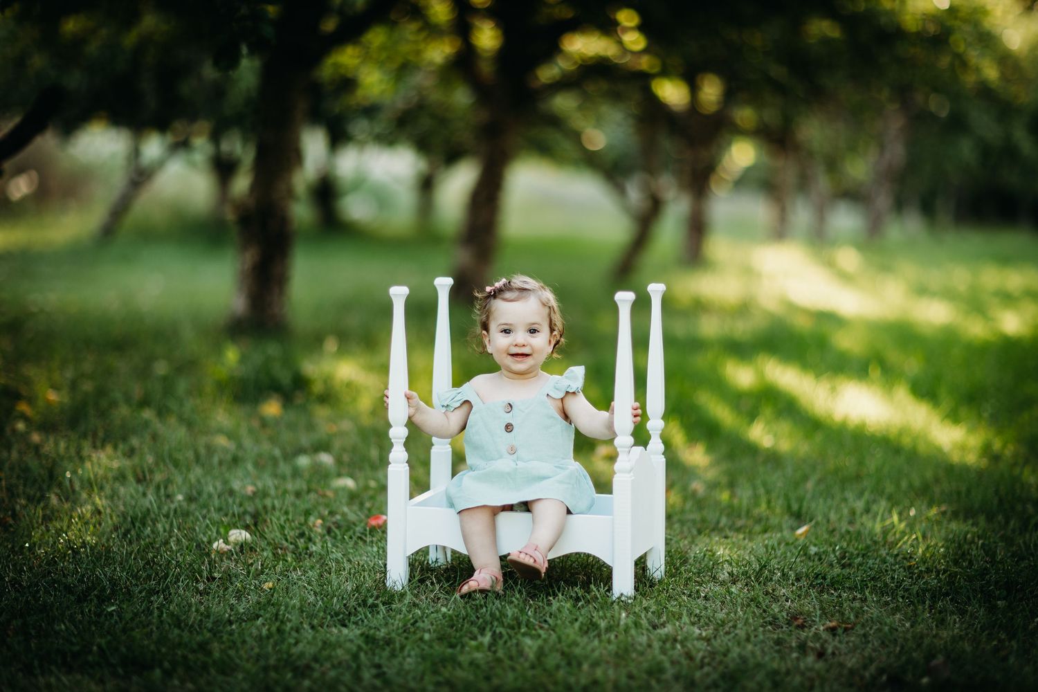 Babyfotograf Moss: En smilende baby som sitter i en hvit dekorativ miniseng utendørs i en grønn eplehage under en barnefotografering