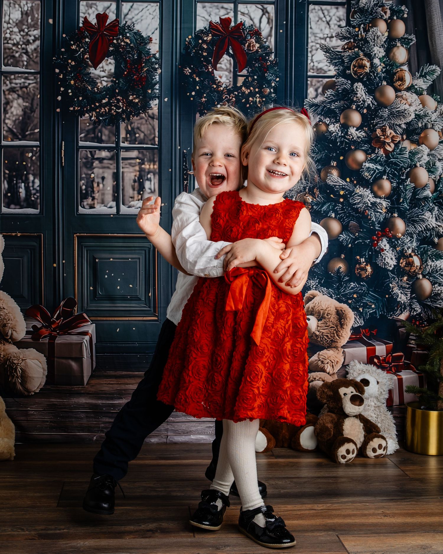 Two happy children in red Christmas outfits hugging and laughing in front of a decorated holiday tree with gifts.
