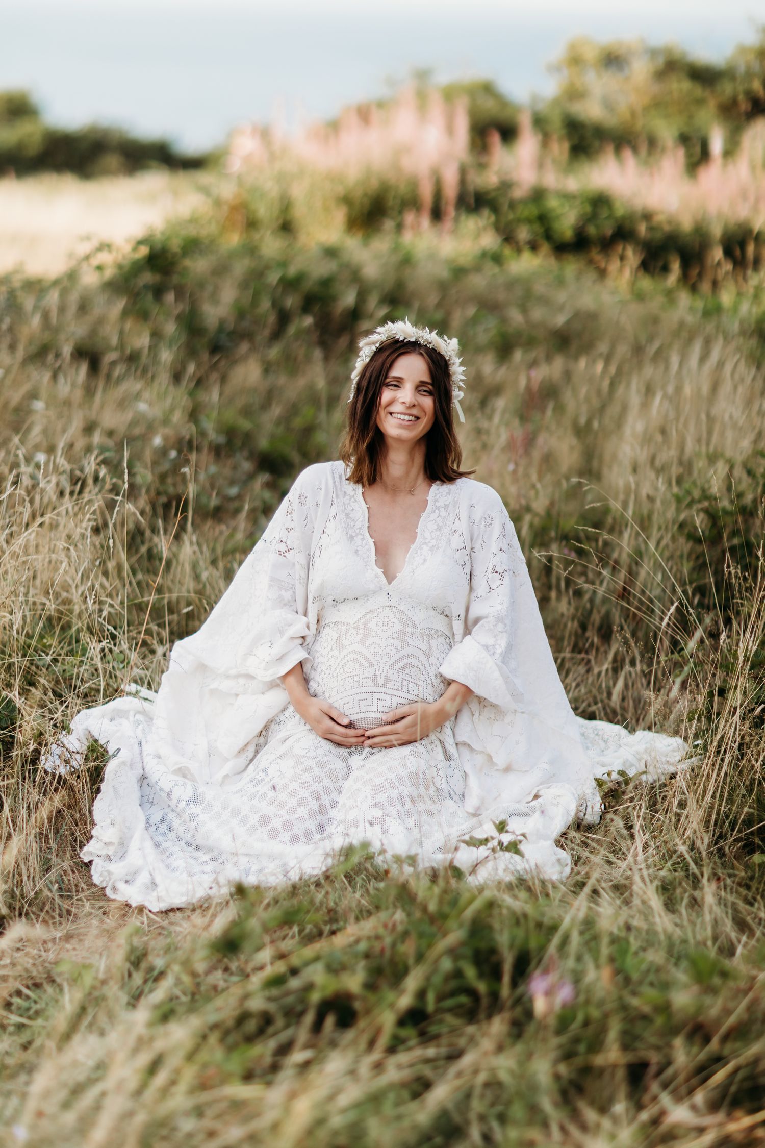 Expectant mother in flowing white lace dress poses gracefully in a sunlit meadow.
