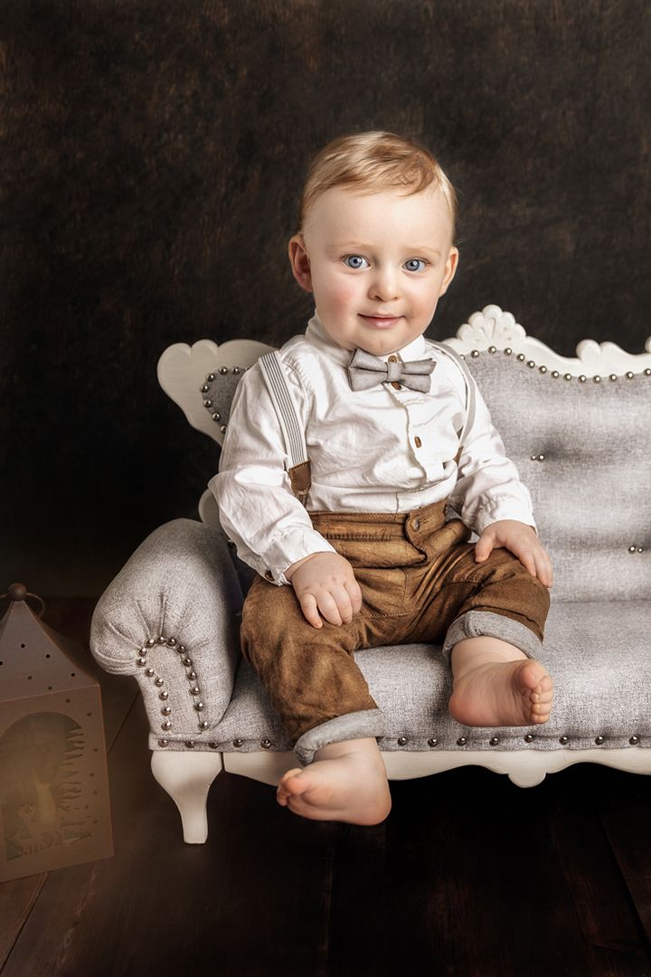 Baby in formal attire sitting on elegant gray tufted couch against dark background.