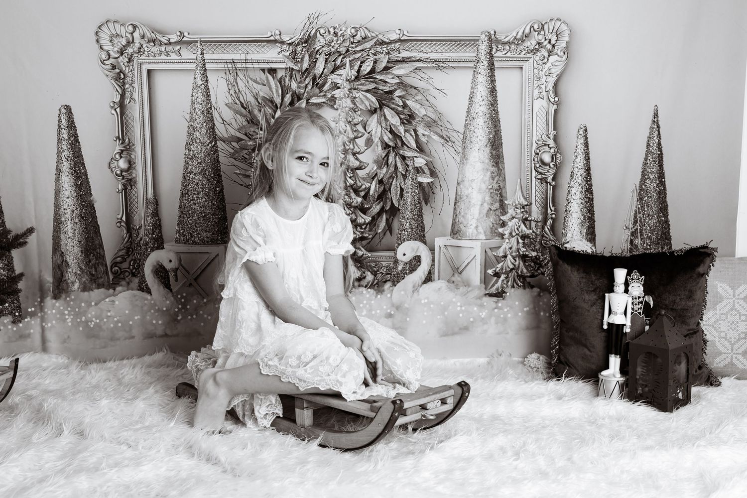 Black and white photo of child sitting on wooden sled with holiday decorations and silver trees.