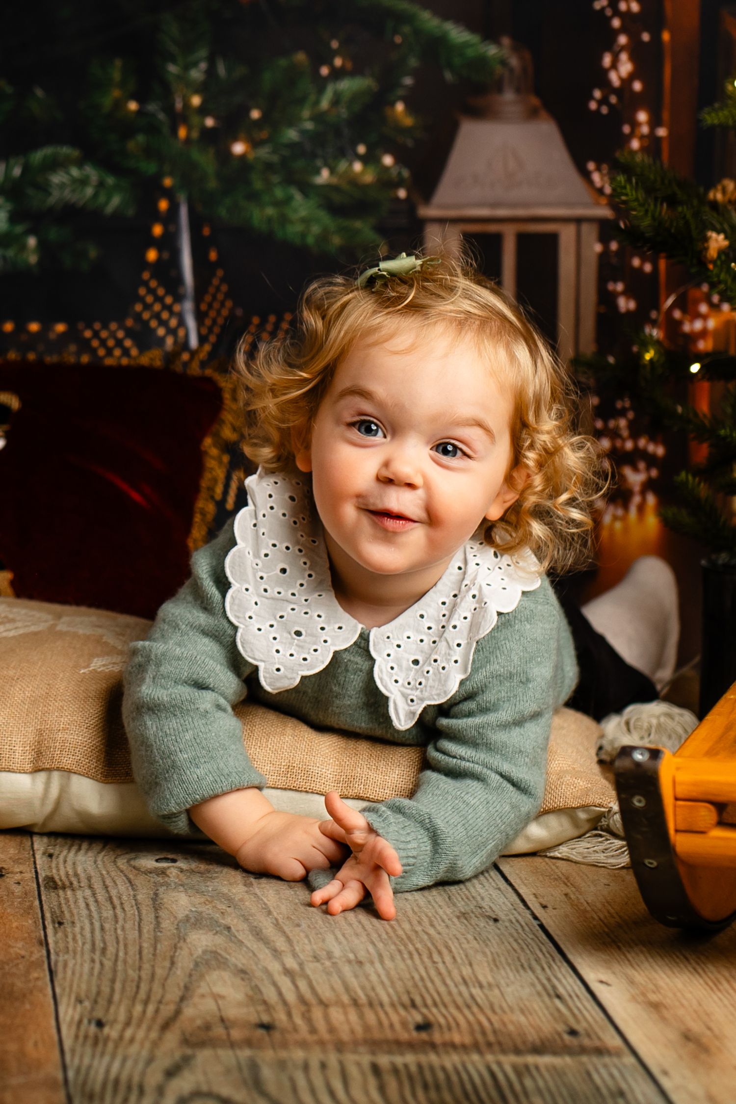 Sweet holiday portrait of toddler in sage green sweater with white collar laying on wooden floor with Christmas lights.